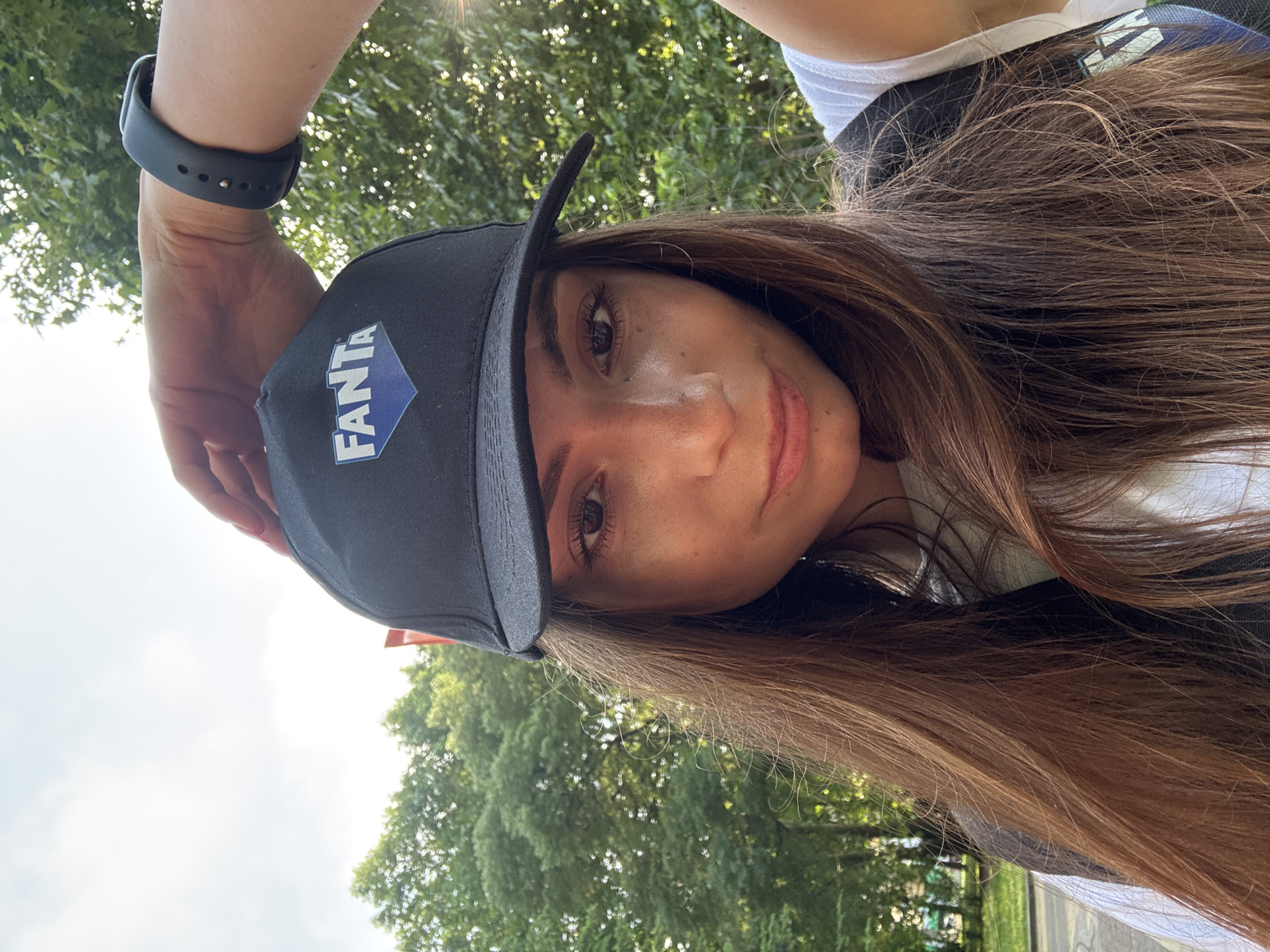A frontal, waist-up selfie depicts a young adult female outdoors. She has long, straight brown hair that cascades over her shoulders and chest. Her face is in focus, with dark eyes gazing directly at the camera and a slight smile on her lips. She wears a black baseball cap with a blue, diamond-shaped logo featuring the white text "FANTA." Her right hand is raised, with fingers resting on the top of the cap, potentially adjusting it. On her left wrist, a dark-colored smart band or fitness tracker with a visible buckle closure is worn. She appears to be wearing a white t-shirt under a dark vest or backpack strap visible on her left shoulder. The background consists predominantly of dense green foliage from trees, suggesting a park or forested area. Bright, diffused sunlight illuminates the scene, creating a lens flare on the right side of the image and a bright, hazy sky above. A paved surface and additional distant greenery are partially visible in the lower-left background. The image is stated to be from the city of Varna, Bulgaria; however, no specific geographic landmarks are discernible within the frame to confirm this.