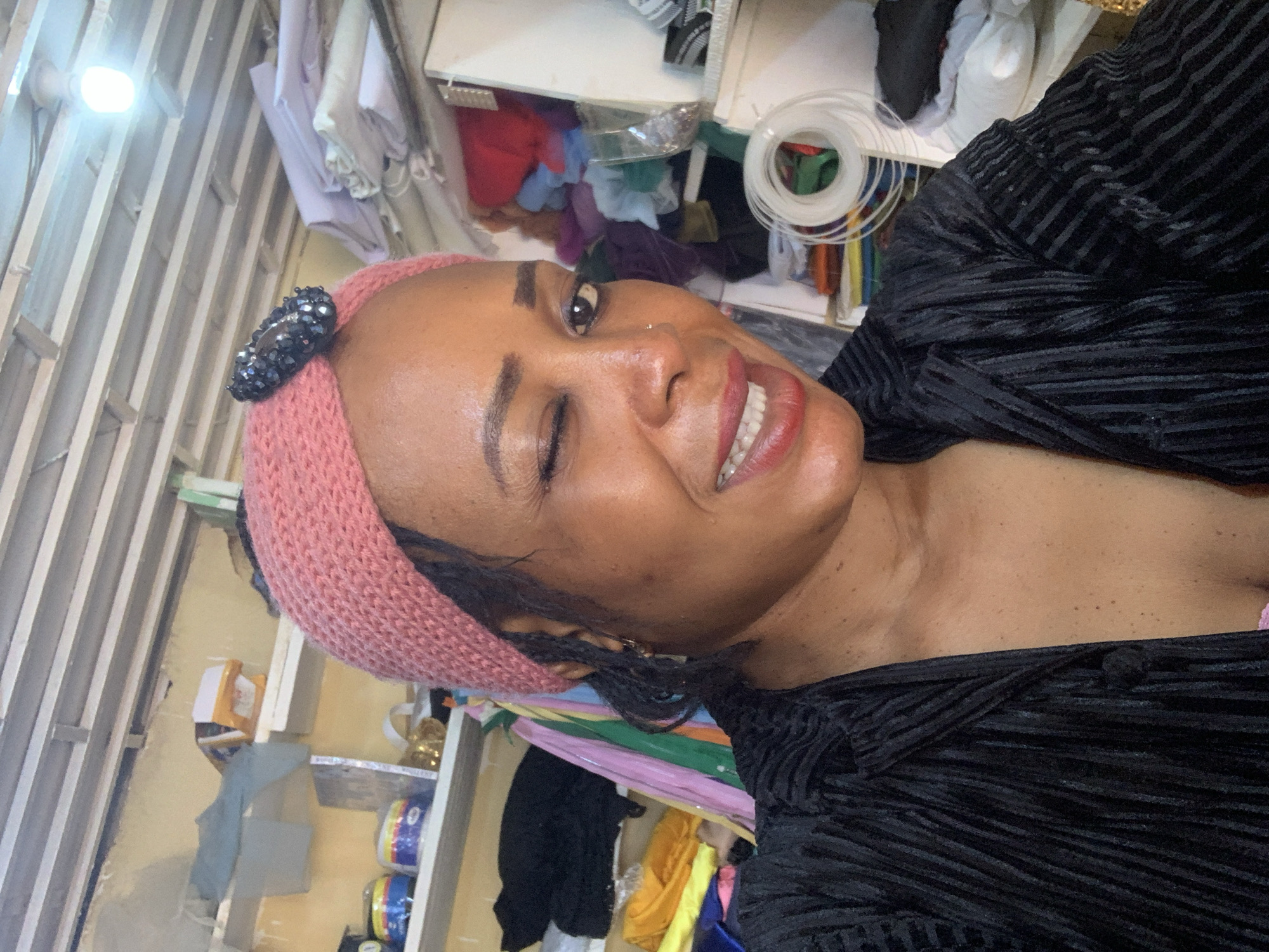 The image shows a close-up, front-facing selfie of a Black woman in what appears to be a workshop or storage area. She is wearing a pink knitted headband with a dark, beaded embellishment and a black, textured, collared shirt. Her right eye is closed in a wink, and her lips are parted, revealing her teeth. The background consists of shelves filled with various items, including fabrics, spools of material, and some packaged goods. The environment suggests a place where materials are stored or processed, possibly a tailor's shop or a craft studio. The specific location within Yelwa, Nigeria, cannot be determined from the image alone.