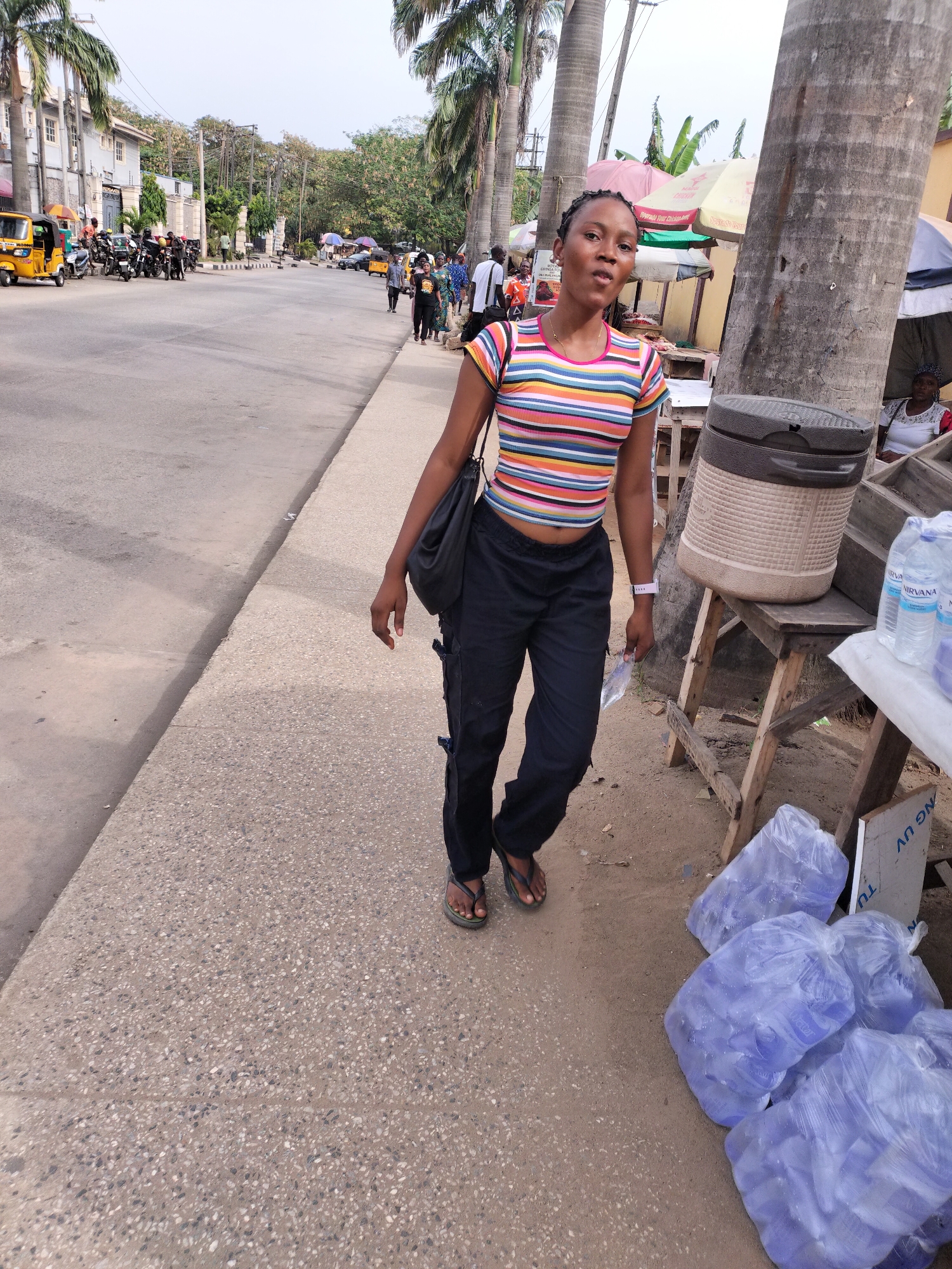 The image captures a street scene in Lagos, Nigeria. An African woman, likely in her late teens or early twenties, is walking on the sidewalk. She wears a short-sleeved, striped crop top and dark-colored pants. She carries a black shoulder bag and wears flip-flops.
The background shows a busy street with motorcycles (okadas) and auto rickshaws (keke napeps) visible. Other people are present on the street and sidewalks. A makeshift stall selling bottled water is situated on the sidewalk next to the woman. Bags of water bottles are stacked near the stall. Palm trees line the street. The overall lighting suggests it is either early morning or late afternoon.
