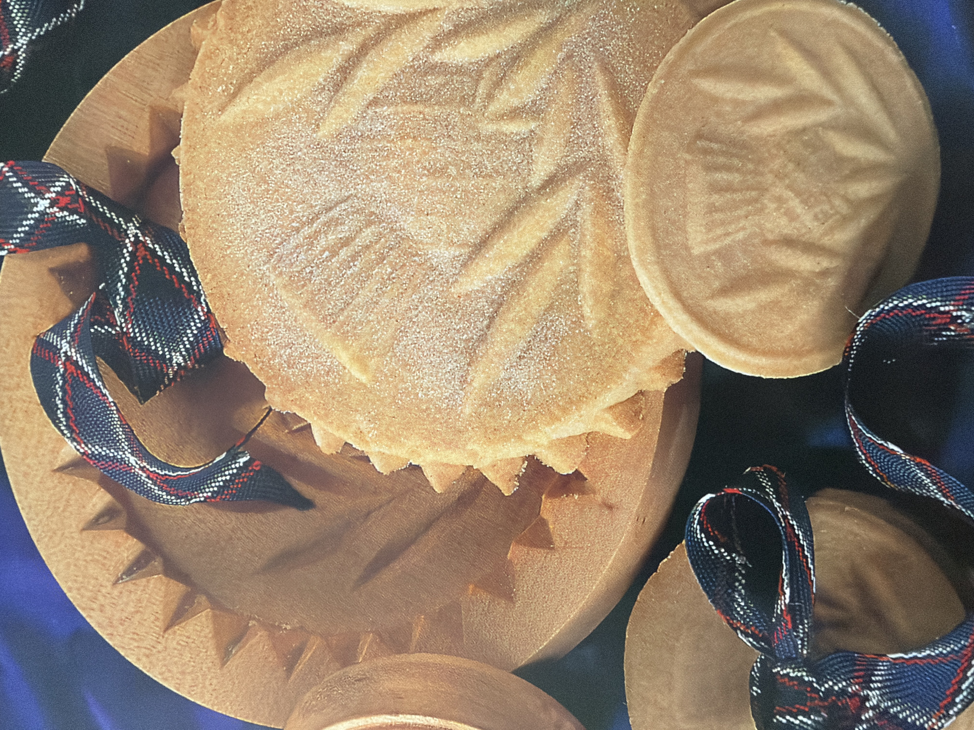 An overhead close-up shot captures a collection of shortbread cookies and a wooden cookie mold. The cookies are round and have intricate patterns pressed into them, resembling leaves or thistles. Some of the cookies are dusted with fine sugar. One cookie is placed on top of the wooden mold, which has a scalloped edge. A tartan ribbon, featuring navy, red, and white plaid, is draped across the cookies and mold. The background is a deep blue, possibly fabric, that is out of focus. The lighting is soft and even, highlighting the textures of the cookies and the wood.