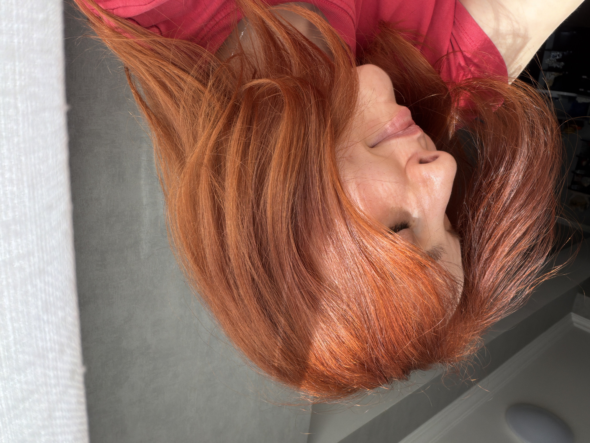 A woman with reddish-orange hair is lying down, possibly indoors. She is wearing a red shirt. The background is out of focus, showing a window with white curtains and a gray wall. There is insufficient information to establish the location as Tbilisi, Georgia.