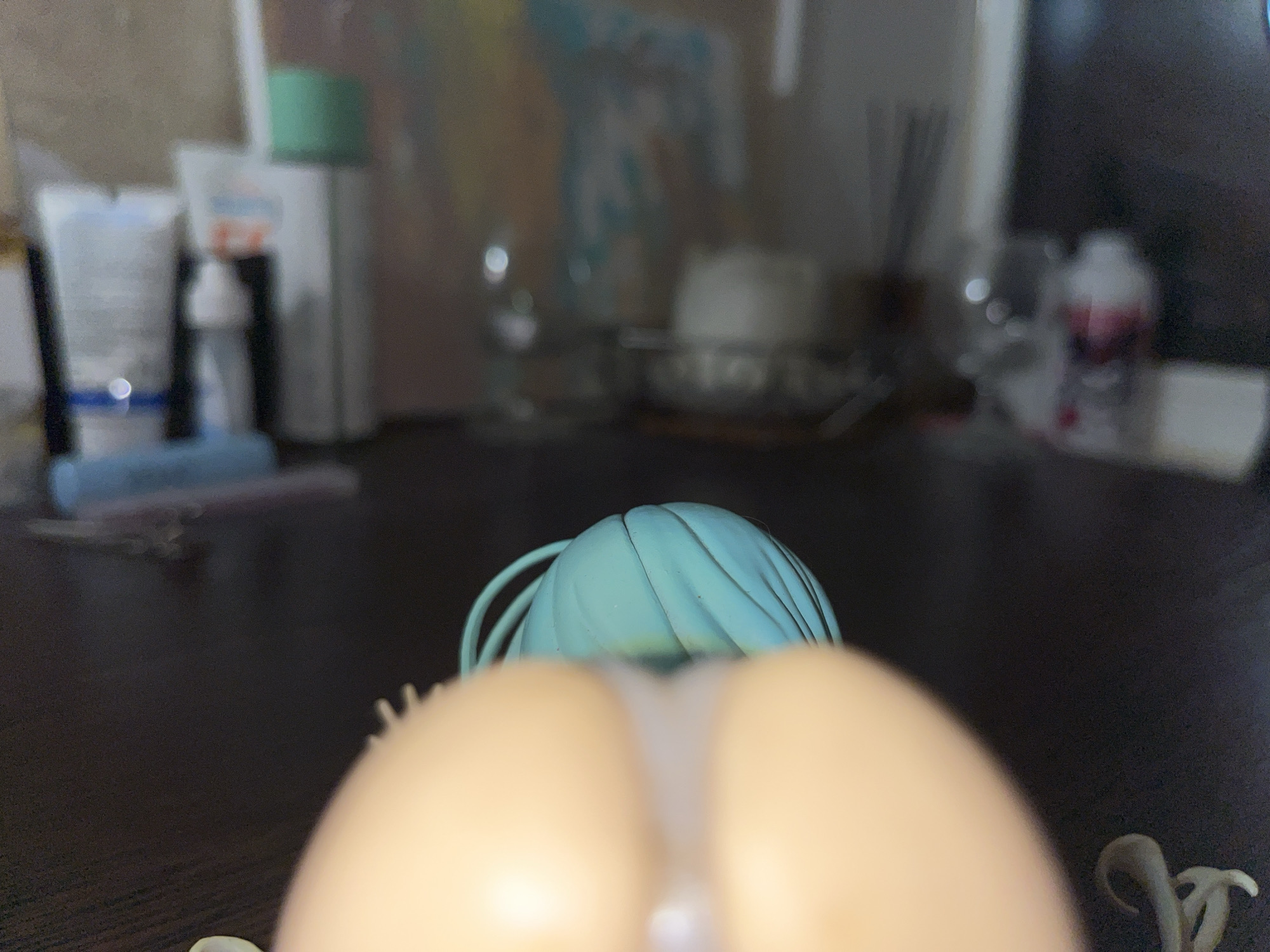 An extreme close-up, slightly angled shot captures the back of a light-skinned doll or figurine with light blue hair. The figurine has two large, rounded, light-skinned buttocks visible in the foreground. Between the buttocks, a sheer white fabric or material is visible, creating a thong-like effect. The background is blurry and consists of a dark wooden surface with various out-of-focus objects including bottles and possibly a painting or poster. The lighting is dim.