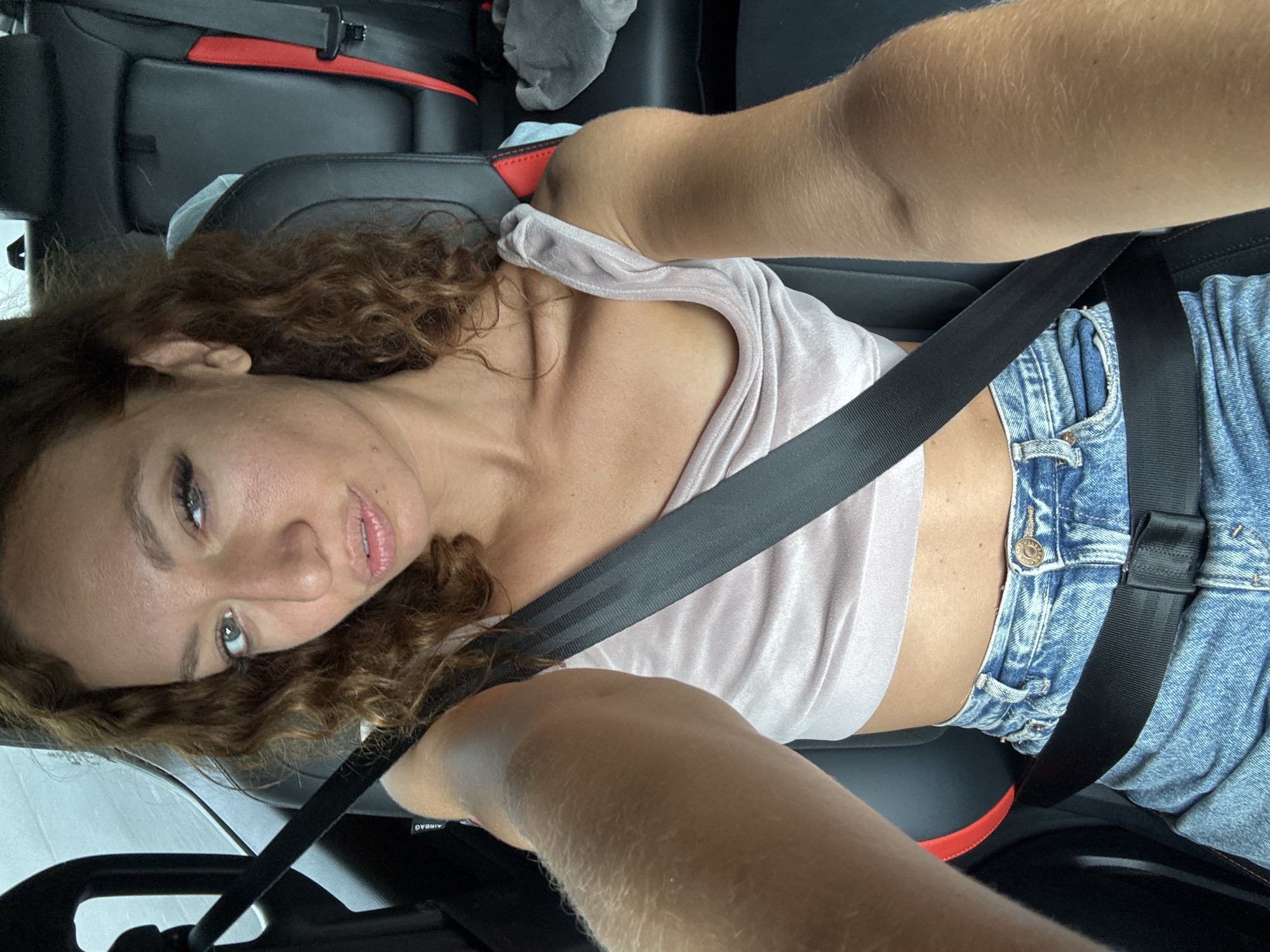 A woman is in a car, wearing a pink top and denim shorts. She is wearing a seatbelt. The location is unknown and cannot be determined from the image.