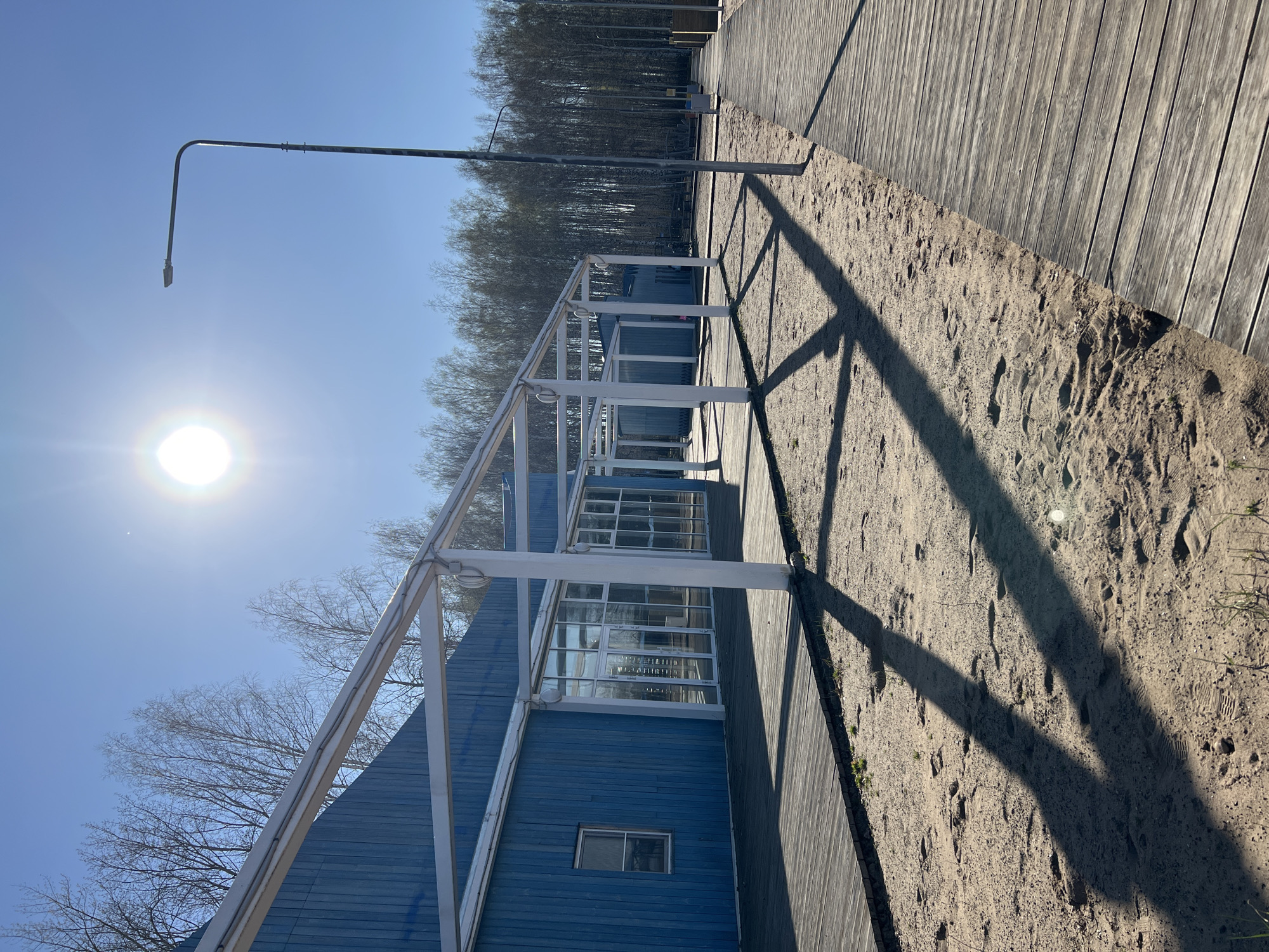 A photograph depicts a blue, A-frame building with a white, open-air enclosure along its side, situated near a boardwalk. The building has multiple windows and one smaller window on the lower level. The boardwalk is constructed of wooden planks and runs parallel to a sandy area. Sunlight is intense, casting sharp shadows of the enclosure onto the sand. A tall, metal lamppost stands in the background. Bare trees are visible behind the building. The photograph appears to have been taken in Chaikovsky, Russia. No people are visible.