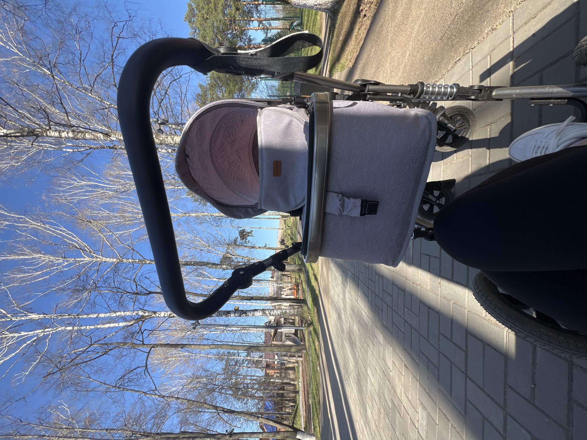 The image shows a view from a stroller on a brick paved path in Votkinsk, Russia. The stroller's gray bassinet is visible, as is the black handle and some of the stroller's chassis. The photographer's legs, wearing dark leggings and a white shoe, are partially visible. To the left, bare birch trees line the path against a blue sky. In the distance, buildings and other trees are visible. The image is taken during daylight hours with the sun casting long shadows on the path.