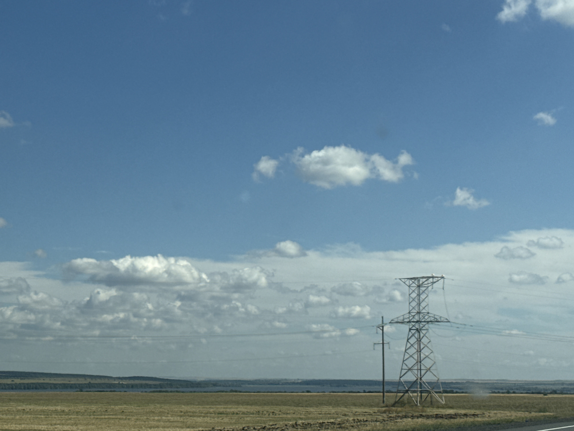 The image depicts a vast, open landscape under a wide expanse of blue sky dotted with white clouds. In the foreground, a dry, golden-brown field stretches out, showing signs of cultivation with visible furrows. A tall, lattice-style electrical pylon stands prominently on the right side of the frame, its metal structure stark against the sky. Another, simpler utility pole is visible to the left of the pylon. In the middle ground, a distant, hazy horizon line separates the field from a body of water, possibly a lake or river, and a low, undulating line of trees. The overall impression is one of a rural, possibly agricultural, setting in a location like Sorochinsk, Russia, characterized by open skies, flat terrain, and essential infrastructure for power transmission.