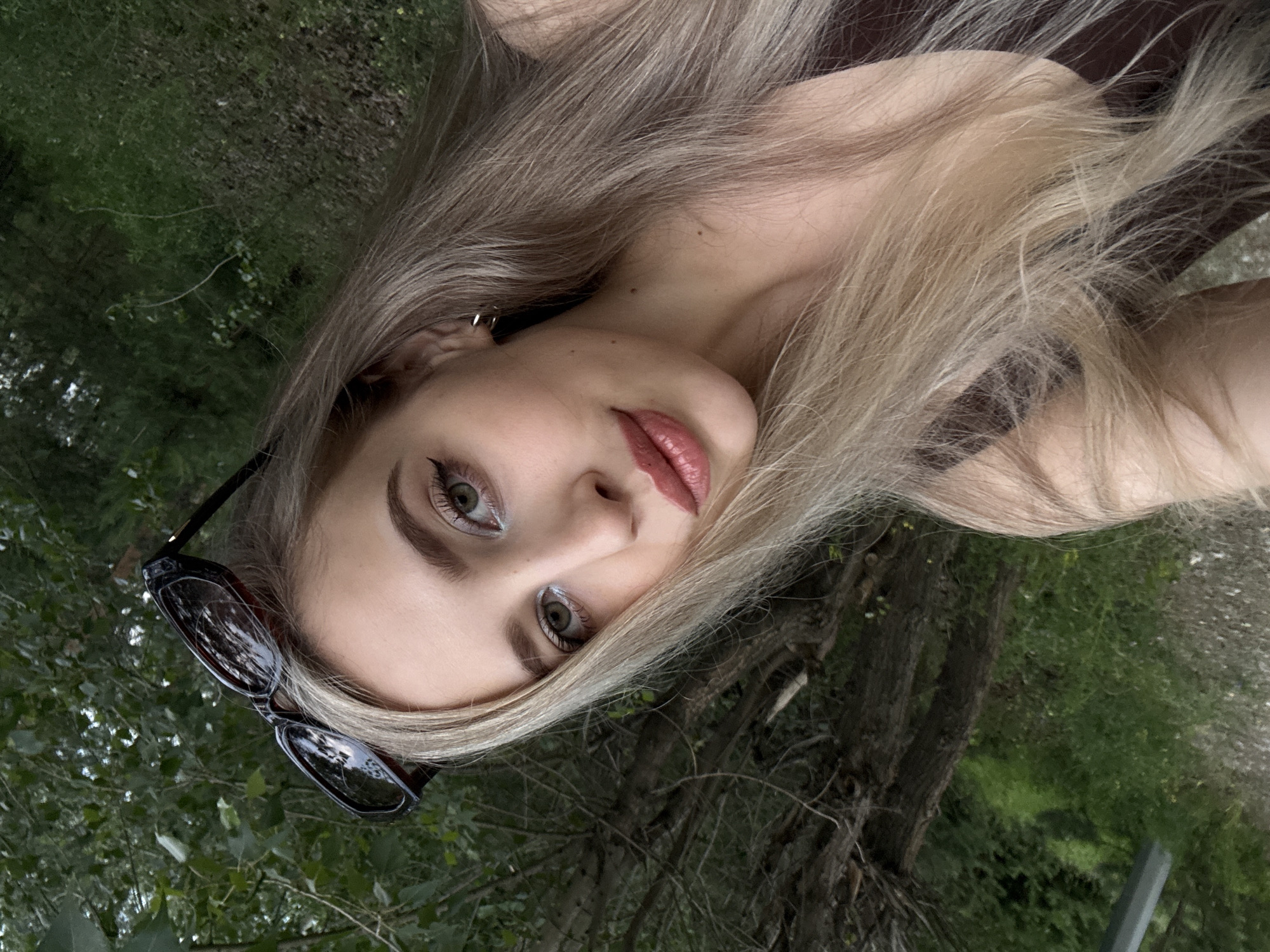 A woman with long blonde hair and light green eyes is looking towards the camera. She is wearing sunglasses on top of her head and makeup, including eyeliner and lipstick. She is wearing a dark brown top. The background is out of focus and appears to be a natural setting with trees and greenery. There is no indication of the location in Krasnoyarsk, Russia.
