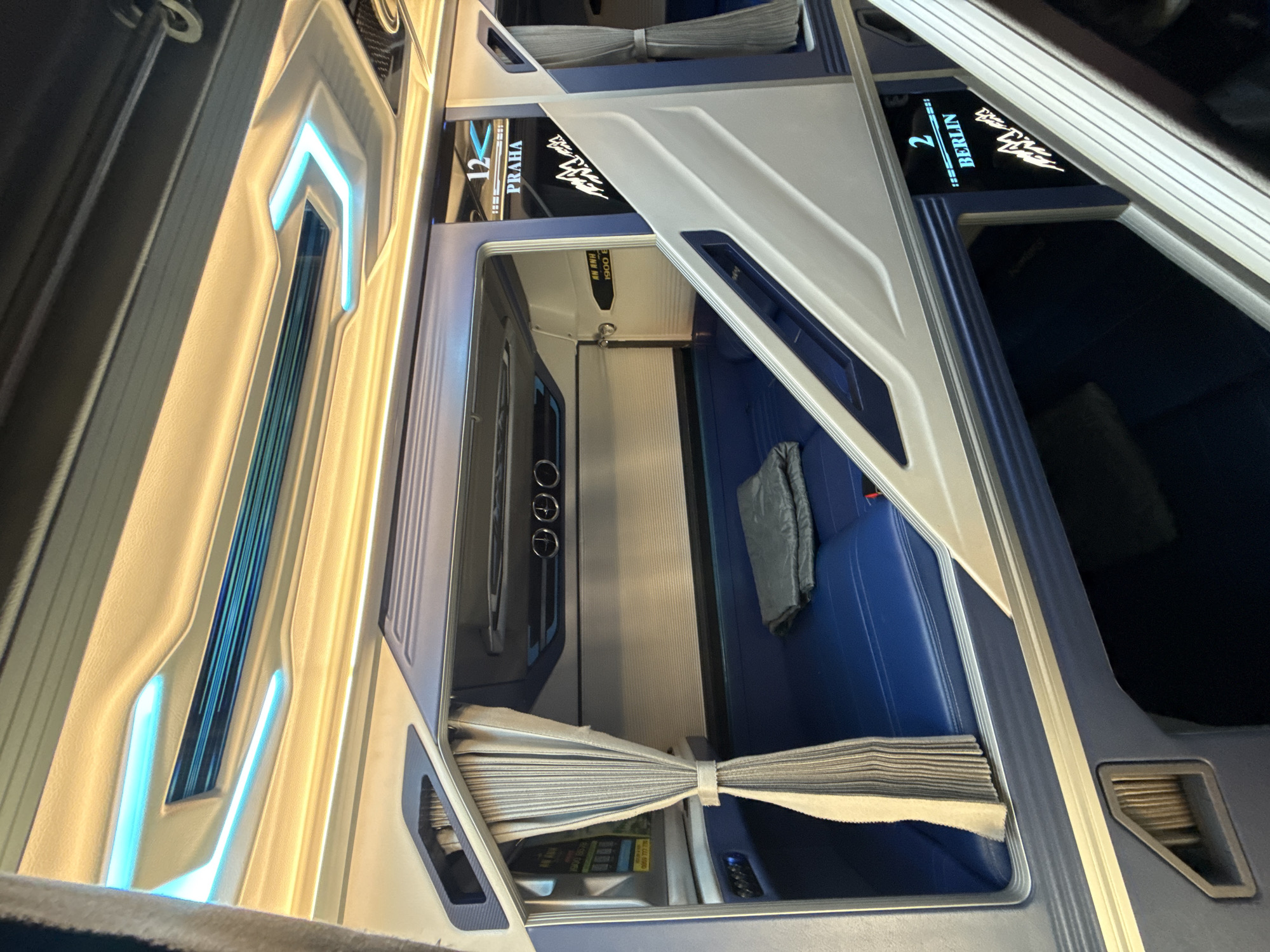The image shows the interior of a sleeper bus cabin, possibly in Da Lat, Vietnam. The cabin features blue upholstered seating, curtains, and modern lighting with blue and yellow accents. Above the sleeping area, there are digital displays indicating "12 PRAHA" and "2 BERLIN," suggesting these are destinations or routes. The overall aesthetic is clean and futuristic, typical of contemporary sleeper bus designs intended for long-distance travel. The lighting creates a moody ambiance within the confined space.