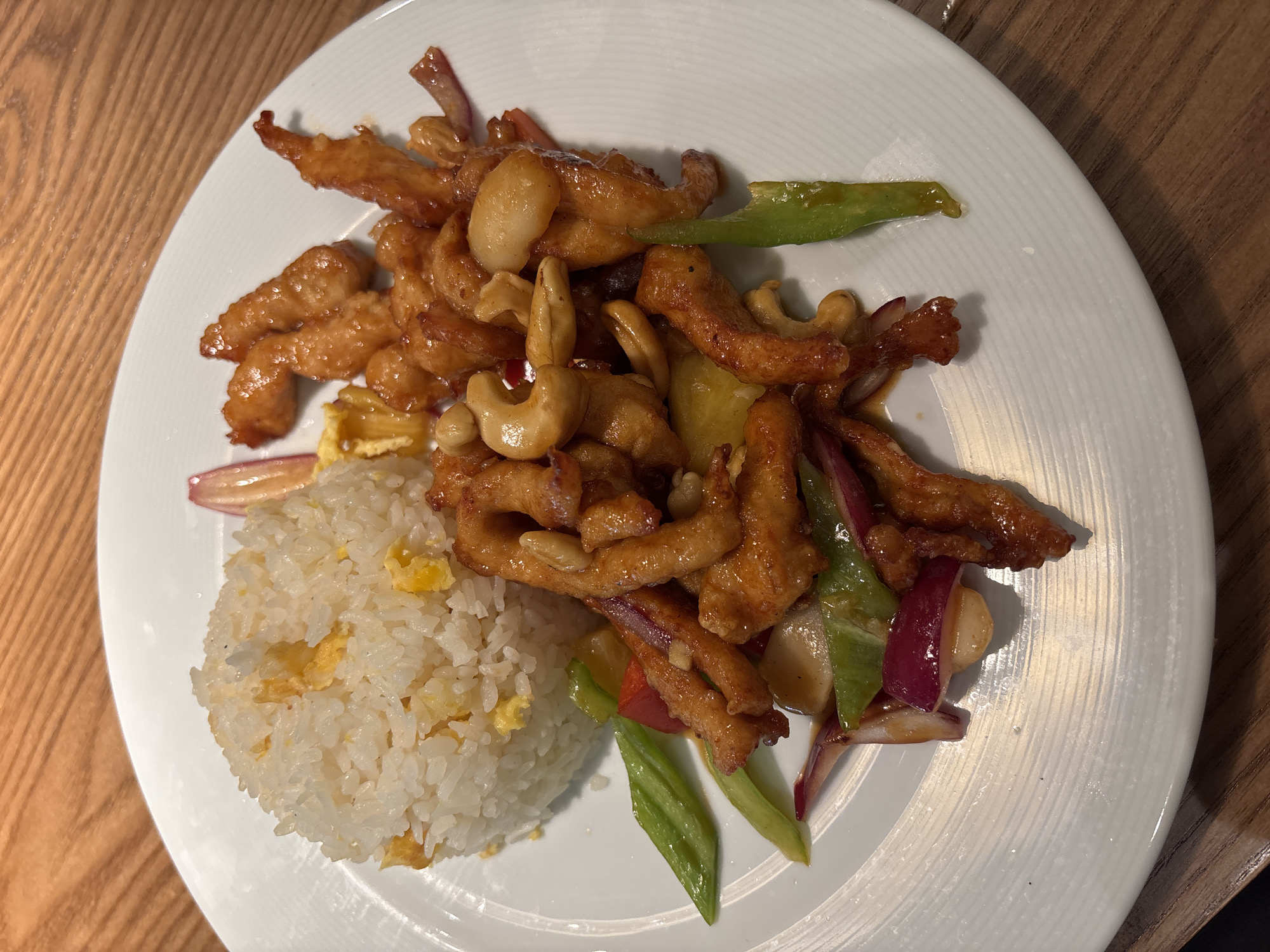This is a top-down, close-up shot of a plate of cashew chicken and rice, presented on a white, circular plate with subtle concentric ridging. The dish itself is the central focus, occupying most of the frame. The cashew chicken is a medley of golden-brown, stir-fried chicken pieces coated in a glossy sauce. Interspersed within the chicken are cashews, slivers of red onion, and pieces of green bell pepper.
To the left of the chicken, a mound of white rice with visible flecks of scrambled egg is nestled. The plate rests on a wooden surface, likely a table, which has a visible grain and a warm, light brown hue. The lighting appears to be natural or well-lit artificial light, casting gentle shadows that highlight the texture of the food and the plate. There is no visible text in the image. The scene suggests a meal being served, possibly in a restaurant or at home. The overall impression is one of a savory and appetizing dish, prepared and ready to be eaten. The image is likely taken indoors, as indicated by the plate and table setting. There are no people visible in the frame. The time of day and weather are not discernible from the image content.