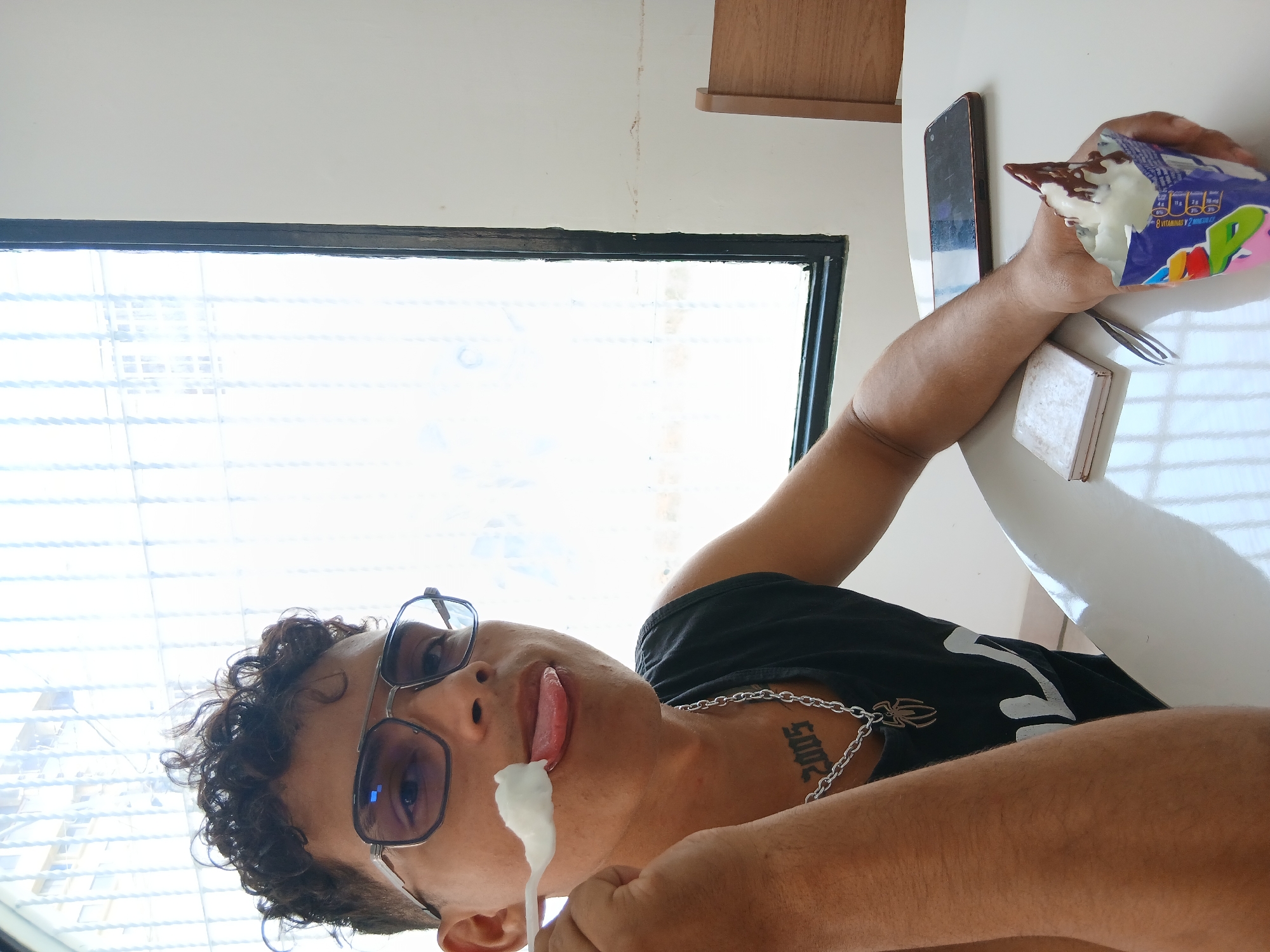 A fair-skinned person with curly dark hair and glasses is seen from a low angle, licking white ice cream from a spoon. The person wears a black t-shirt and a silver chain necklace, and a tattoo with the number "2005" is visible on their neck. The background shows a window with a view of a building and a white ceiling. On a white table, there is a smartphone, a white tile, a fork, and the remainder of a partially eaten ice cream in a colorful wrapper. The location is Juan Griego, Venezuela.