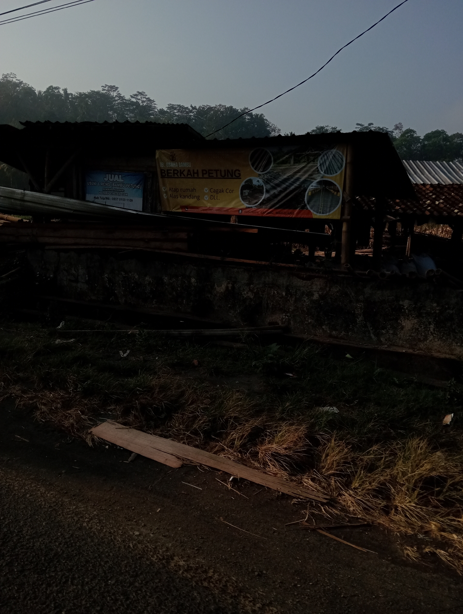 The image depicts an outdoor scene with a hazy sky and lush green hills in the background. In the foreground, a rustic structure with a dark corrugated roof is visible. A banner with text in Indonesian hangs from the structure, advertising a business called "BERKAH PETUNG" which sells "USUK & RENG (BAMBU PETUNG)". The banner also lists various products and services offered, including roofing, cage flooring, and other items. Below the structure, there is a rough wall and a pile of what appears to be bamboo or wooden poles. The ground in front of the structure is covered with dry grass and debris, and a section of a dirt road is visible. The scene appears to be located in Krajan Kidul, Indonesia, as indicated by the text on the banner. The overall impression is that of a rural or semi-rural area, possibly a small business operating in a natural setting.