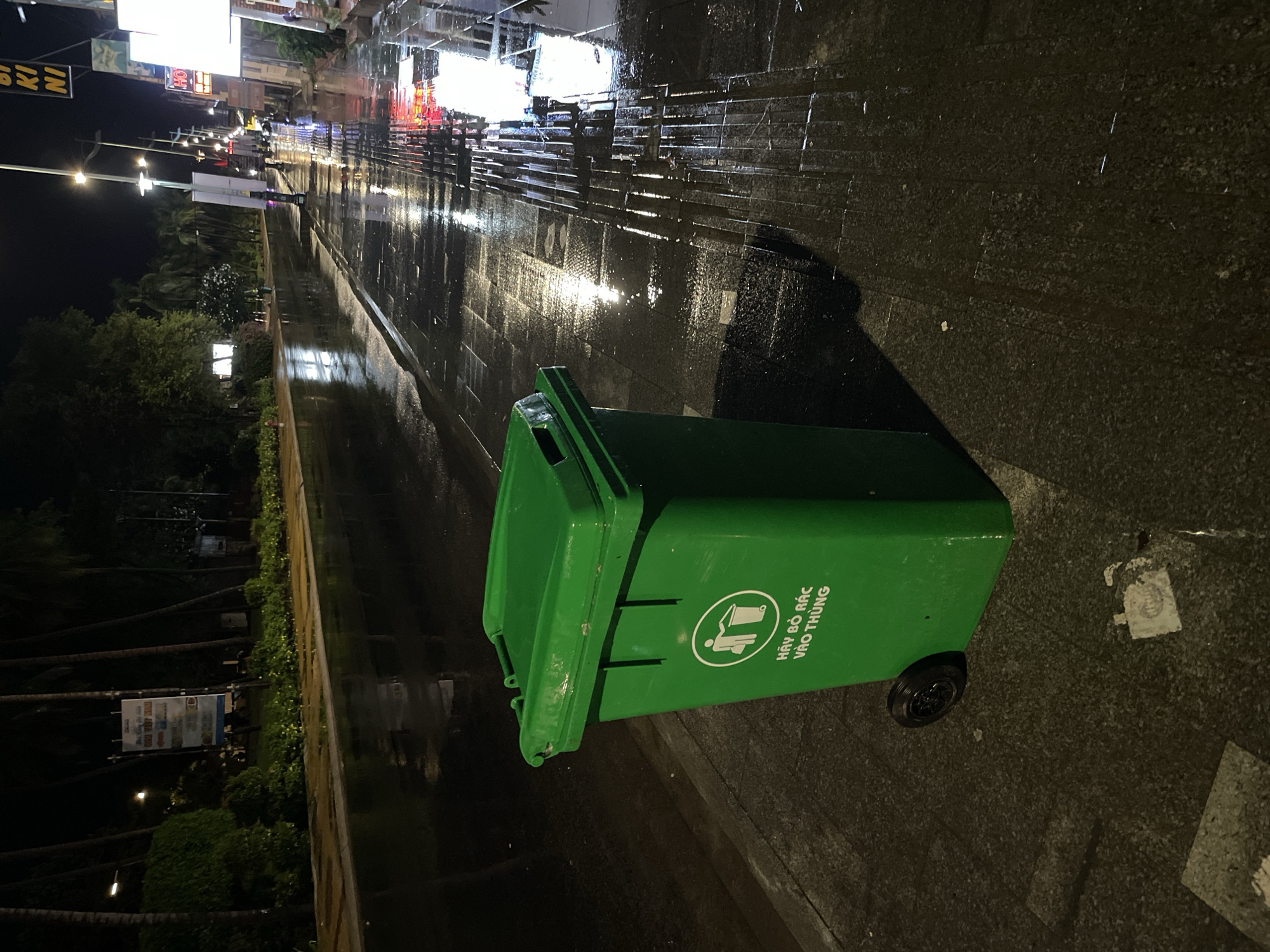 This night-time photograph, taken in Phu Thuy Ward, Vietnam, captures a wet urban street scene, likely a sidewalk or pedestrian area, after rainfall. The main subject in the foreground is a large, bright green plastic wheeled trash bin. Its lid is slightly ajar. On the side of the bin, there is a white circular symbol depicting a person disposing of an item into a bin, accompanied by the Vietnamese text "HÃY BỎ RÁC VÀO THÙNG," which translates to "PLEASE PUT TRASH INTO THE BIN."
The ground is dark and highly reflective, showing numerous elongated streaks and patches of light, indicating it is wet from recent rain. In the background, a street or promenade stretches into the distance, lined with streetlights, brightly lit commercial buildings, and dense foliage including palm trees. The artificial lights from the establishments and streetlamps cast vibrant reflections on the wet surface, creating a shimmering effect. Partially visible signs on distant buildings include one with the text "BI KI NI" and another with "HOT." No people or specific activities are visible in the static scene. The dark sky confirms the time of day as night, illuminated by the urban lights.