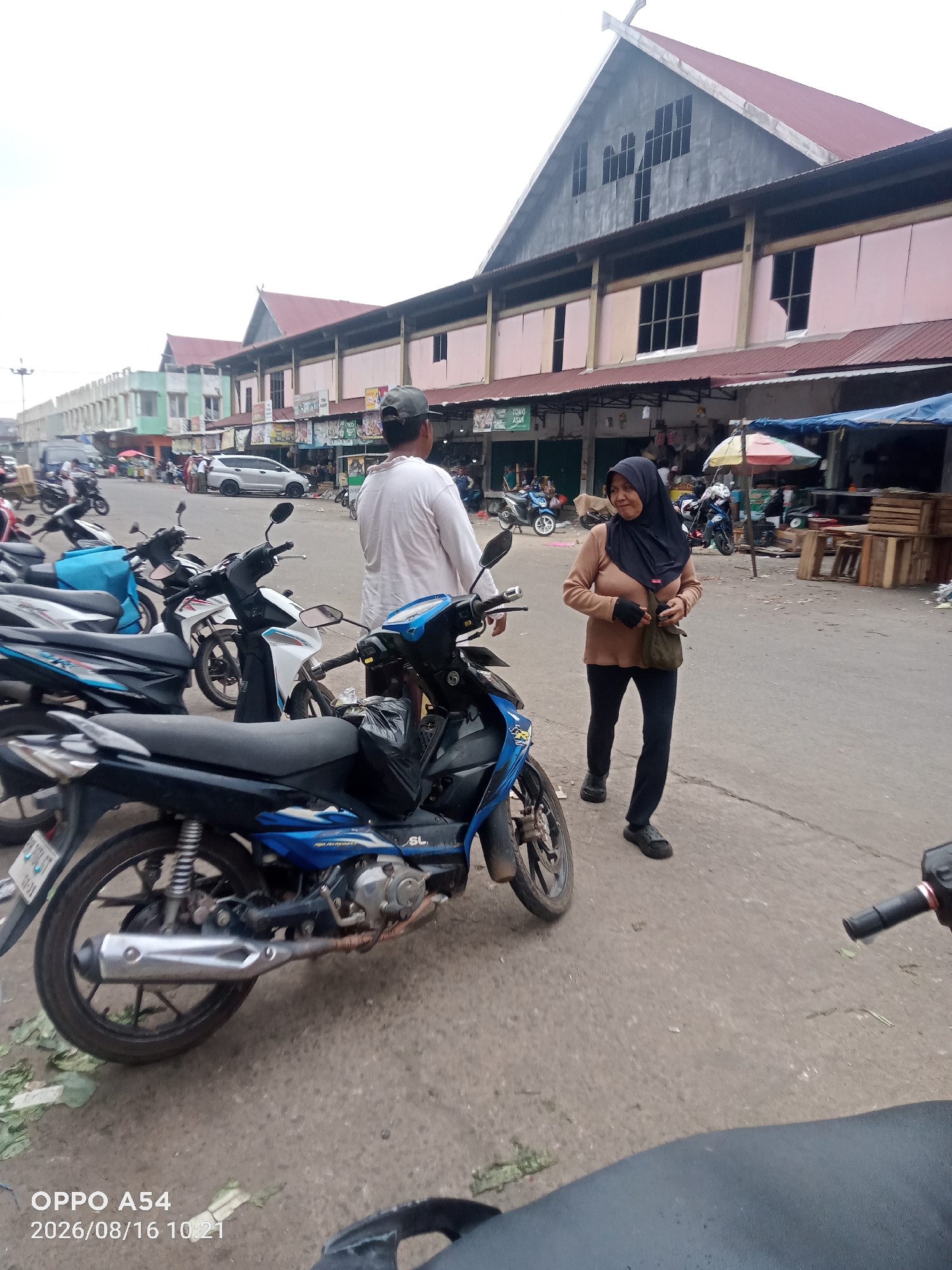 The image shows an outdoor scene in a market area, likely in Jambi, Indonesia. In the foreground, several motorcycles are parked. A blue and black motorcycle is prominently displayed. To the right, a woman wearing a dark hijab and a light brown long-sleeved shirt and dark pants walks away from the viewer. She is carrying a dark green bag over her shoulder and appears to be wearing dark slip-on shoes. Behind her, a man wearing a white long-sleeved shirt and a dark baseball cap stands facing away from the camera. In the background, there is a long building with a red roof and light pink walls, featuring several windows. A colorful umbrella is visible near a stall, and wooden crates are stacked. Various other parked motorcycles and people can be seen further in the background, suggesting a busy marketplace. The ground is paved with asphalt, and some debris, including green leaves, is scattered on it. The sky is overcast. The bottom left corner of the image displays "OPPO A54 2026/08/16 10:21".