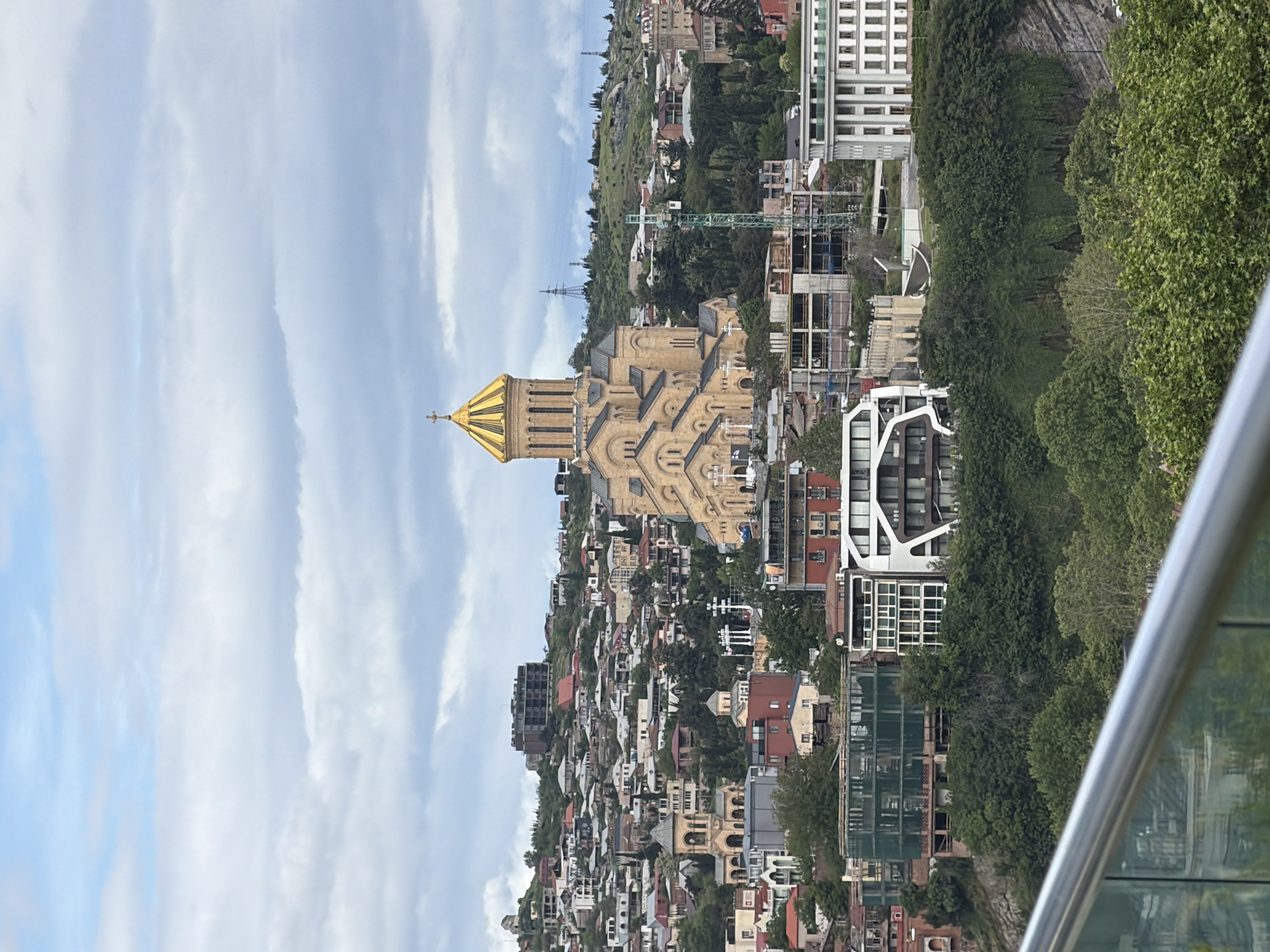 The image is an outdoor shot of the Holy Trinity Cathedral of Tbilisi, Georgia. The cathedral's golden dome is visible, along with its detailed facade. The background shows a cluster of buildings on a hillside, part of the urban landscape of Tbilisi. A construction crane is also visible. Trees are scattered throughout the scene, adding greenery to the urban environment. A part of a glass barrier or balcony is seen in the foreground. The sky is overcast with clouds.