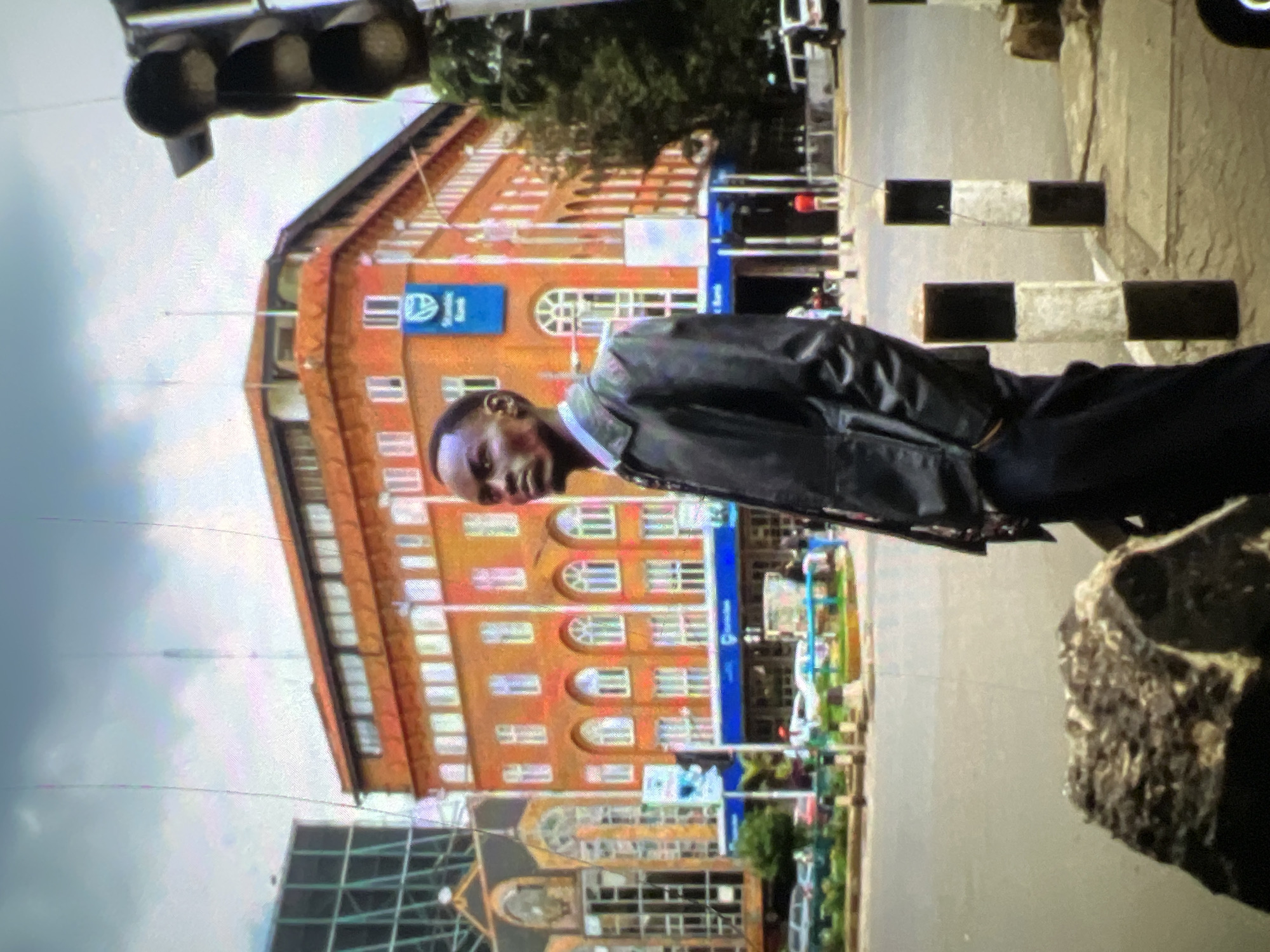 A medium shot captures a young Black man standing on a city street, facing right and looking off into the distance. He is wearing a dark leather jacket over a light collared shirt and dark trousers. The man is positioned in the foreground, slightly to the left of center, with a blurred grey rock in the lower left corner of the frame.
Behind him, a prominent orange brick building with arched windows occupies the background. A blue banner with a white logo and text, appearing to be an advertisement for a bank, is visible on the side of this building. To the left of the orange building is a modern glass-fronted building. The street scene includes vehicles, traffic lights, and bollards. The sky is overcast with some visible lines on the image that may be artifacts from the capture or display. The overall setting appears to be an urban environment, likely a commercial district.