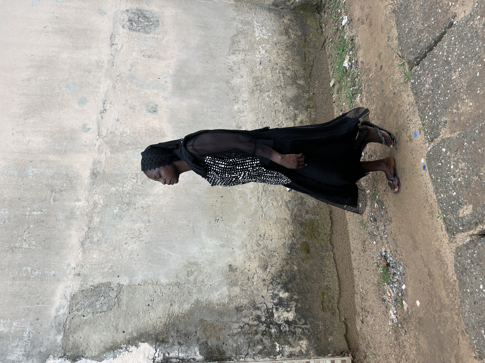 A woman walks from left to right in profile, facing the right side of the frame. She is wearing a black headscarf, a black and white polka dot blouse with sheer sleeves, and a long black skirt. She has dark skin and her eyes are closed. She is wearing dark flip-flops. The woman is walking on a dirt path that has some gravel and debris. To the right of the path is a cracked concrete curb and sidewalk. Behind the woman is a textured, weathered concrete wall with areas of dark staining and lighter patches. The scene appears to be an outdoor environment, possibly a street or alleyway in Abuja, Nigeria.