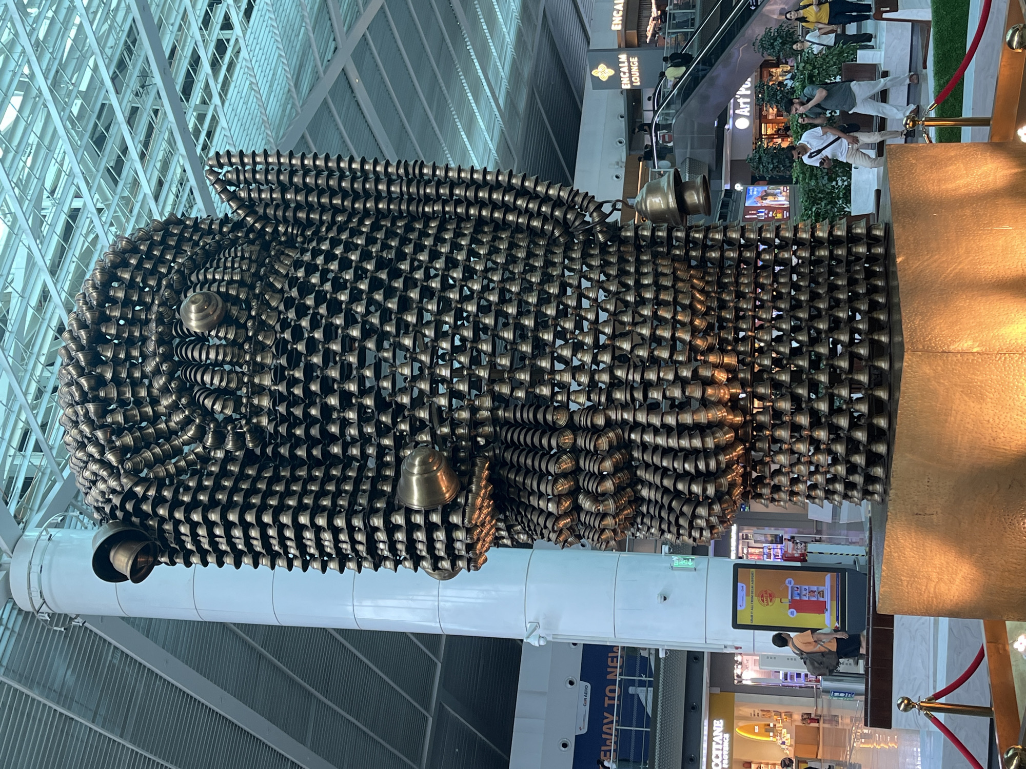 The image depicts a large, intricate sculpture made of many small, brass bells, forming a stylized human head or figure. The sculpture is located indoors, likely within an airport or a large public building, indicated by the modern architecture, signage, and presence of escalators and shops in the background. Specifically, the signage "ENCLAM LOUNGE" and "Art P" suggests this could be Indira Gandhi International Airport in Delhi, India. The sculpture is positioned on a golden pedestal. Several people are visible in the background, some walking and others standing, with some engaging in photography. The environment appears to be well-lit, with a high ceiling and metallic structural elements. A person with a backpack is standing near a digital display screen to the left of the sculpture.