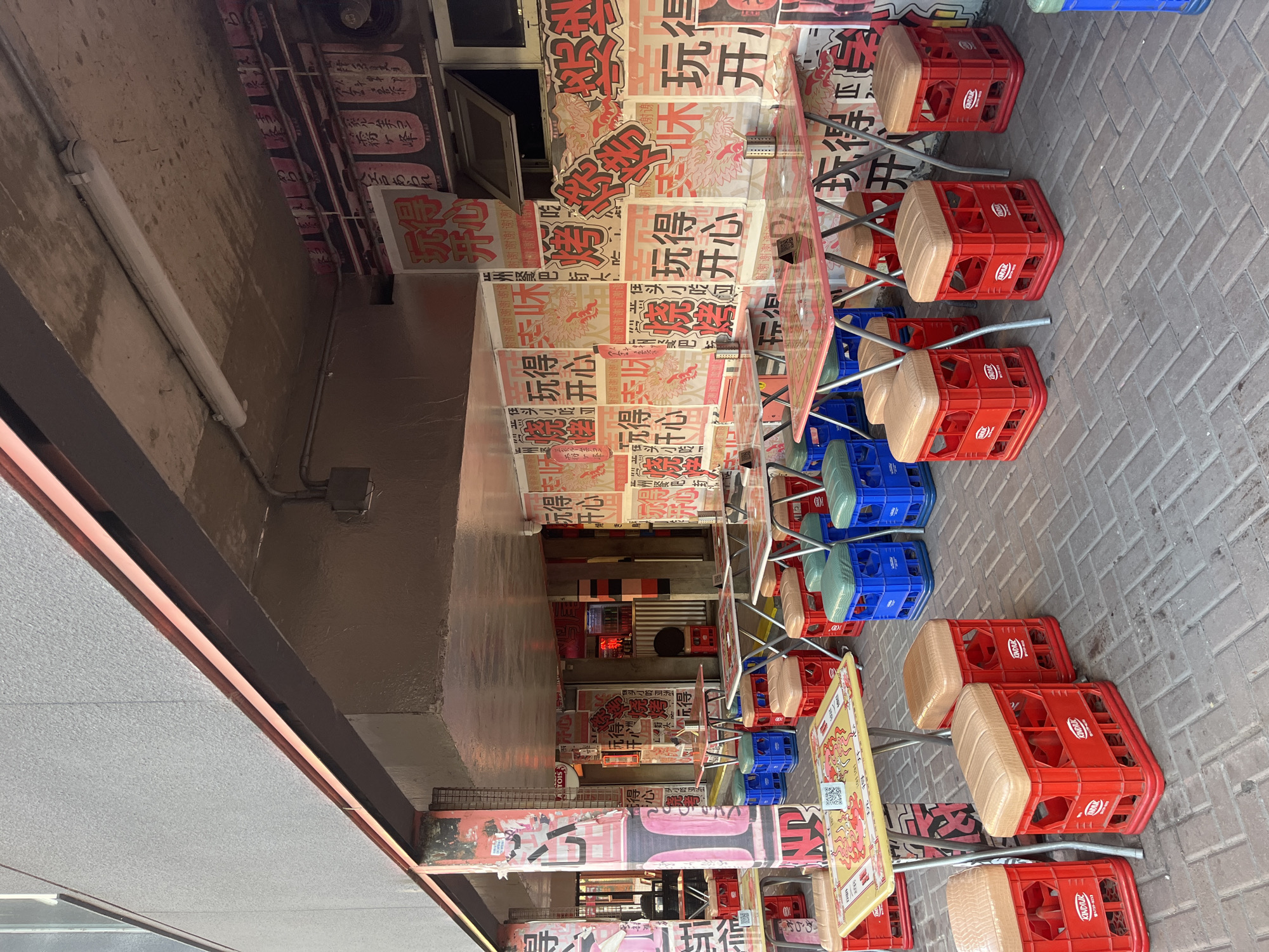 The image captures a narrow alleyway in a bustling area. The alley walls are covered in signs, likely advertisements or promotional materials in a non-English language. Tables and chairs line the passageway, suggesting an outdoor dining or gathering space. The chairs are made from plastic crates of red and blue colors with a tan cushion on top. The alleyway floor is paved with light grey rectangular tiles. The lighting appears to be a mix of natural daylight filtering through the alley's opening and artificial lights from the nearby establishments. Based on the signs, the location could be a restaurant or food stall area. The image does not provide specific visual evidence that would allow for definite confirmation of its location in Buenos Aires, Argentina.