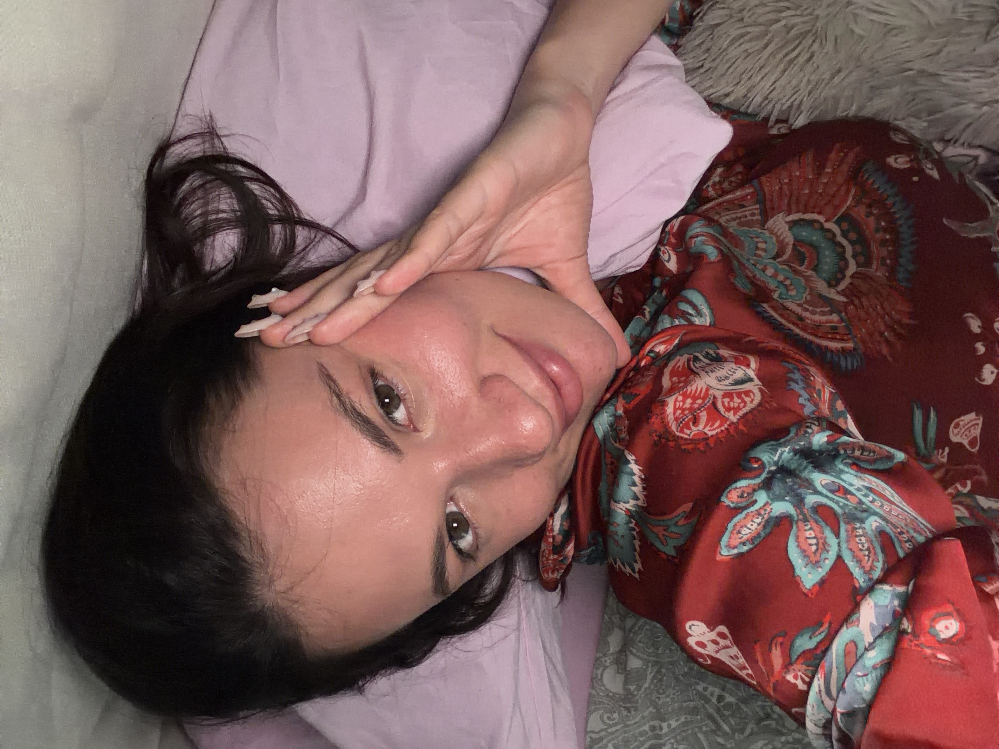 A woman with dark hair is lying in bed, looking at the camera. She is wearing a red patterned pajama top and has her hand resting on her face. The background includes a pink pillow and a grey textured blanket. The scene appears to be indoors.