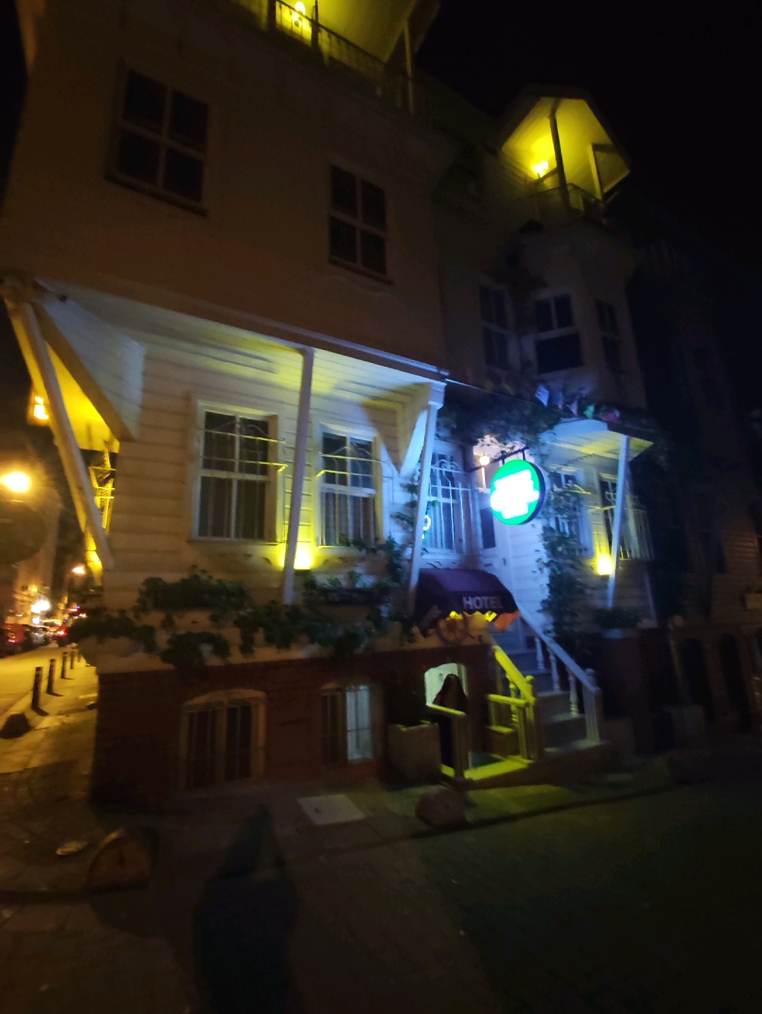 The image captures a nighttime street scene in Kadıköy, Turkey, focusing on the facade of a three-story building that appears to be a hotel. The building is illuminated by warm yellow lights, highlighting its white wooden exterior with dark window frames. Multiple windows with visible interior lights are present on each floor. A prominent green circular sign with white lettering, likely indicating "Hotel," is mounted on the second story, accompanied by a red awning over an entrance. The ground level features a brick base with several windows and planters overflowing with greenery. A staircase with yellow railings leads to an upper entrance. The street in front of the building is paved, with bollards lining the side. A few vehicles are visible in the background, suggesting a relatively busy urban environment. A dark figure is partially visible in the lower right quadrant of the image.