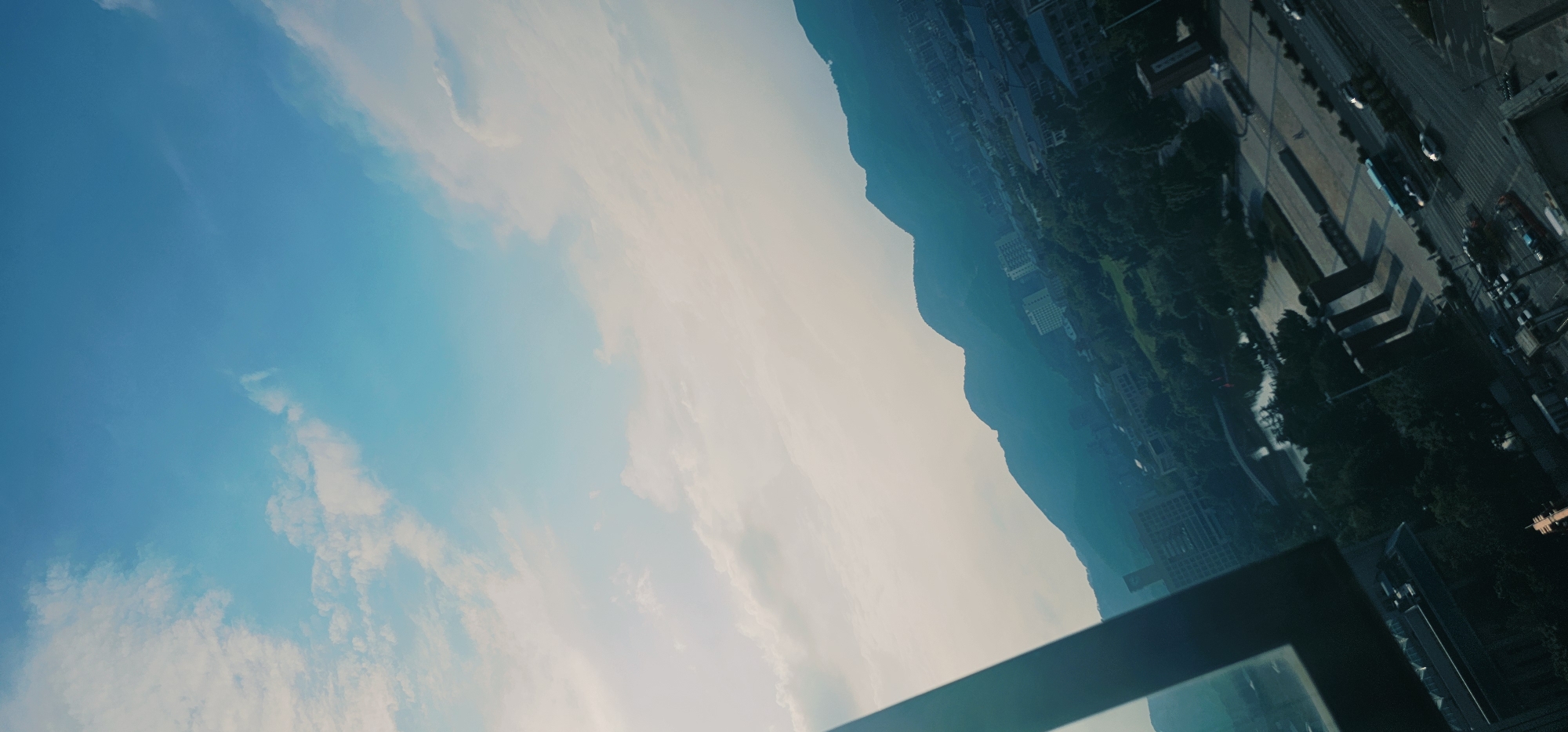 The image is an angled aerial view of a city with mountains in the background. In the foreground, a dark, angular structure, possibly a window frame or balcony railing, occupies the left side of the frame. Beyond this, a landscape unfolds with buildings, green trees, a body of water, and roads. The buildings are varied in size and design, with some appearing to be multi-story residential or office blocks. The roads show some traffic, including cars and a bus. In the distance, a range of mountains stretches across the horizon, silhouetted against a sky with scattered clouds. The overall impression is one of a semi-urban environment bordering a natural mountainous landscape, likely taken from a high vantage point such as a tall building. The location is the Chongqing High-tech Zone, China.