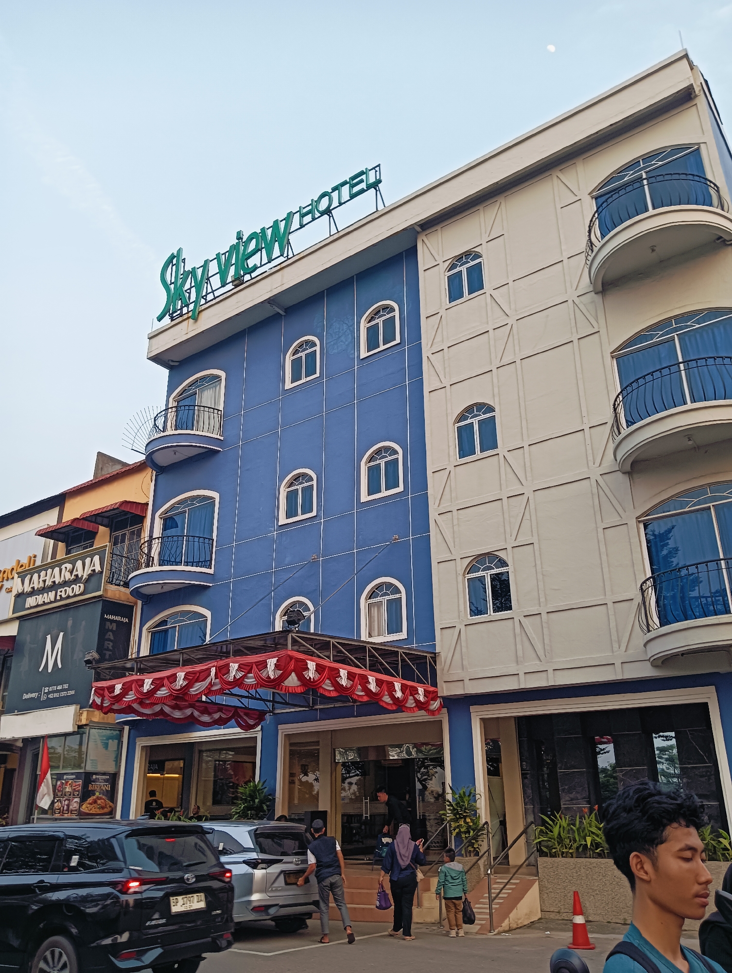 The image shows the Skyview Hotel in Batam, Indonesia. The hotel is a multi-story building with a blue facade on the left and a beige facade on the right. Several arched windows are visible on both sides of the building, some with balconies. The hotel's name, "Skyview HOTEL," is displayed in green lettering on the top of the building.
In front of the hotel, there is an awning with red and white fabric decorations. Below the awning are the entrances to the hotel and an adjacent establishment. To the left of the hotel, another building is visible with signage that reads "MAHARAJA INDIAN FOOD."
Several cars are parked on the street in front of the establishments, including a black SUV and a silver SUV. A few people are walking in the foreground and near the entrances. The sky is overcast. The overall scene depicts a street-level view of a hotel and its surroundings in an urban setting.