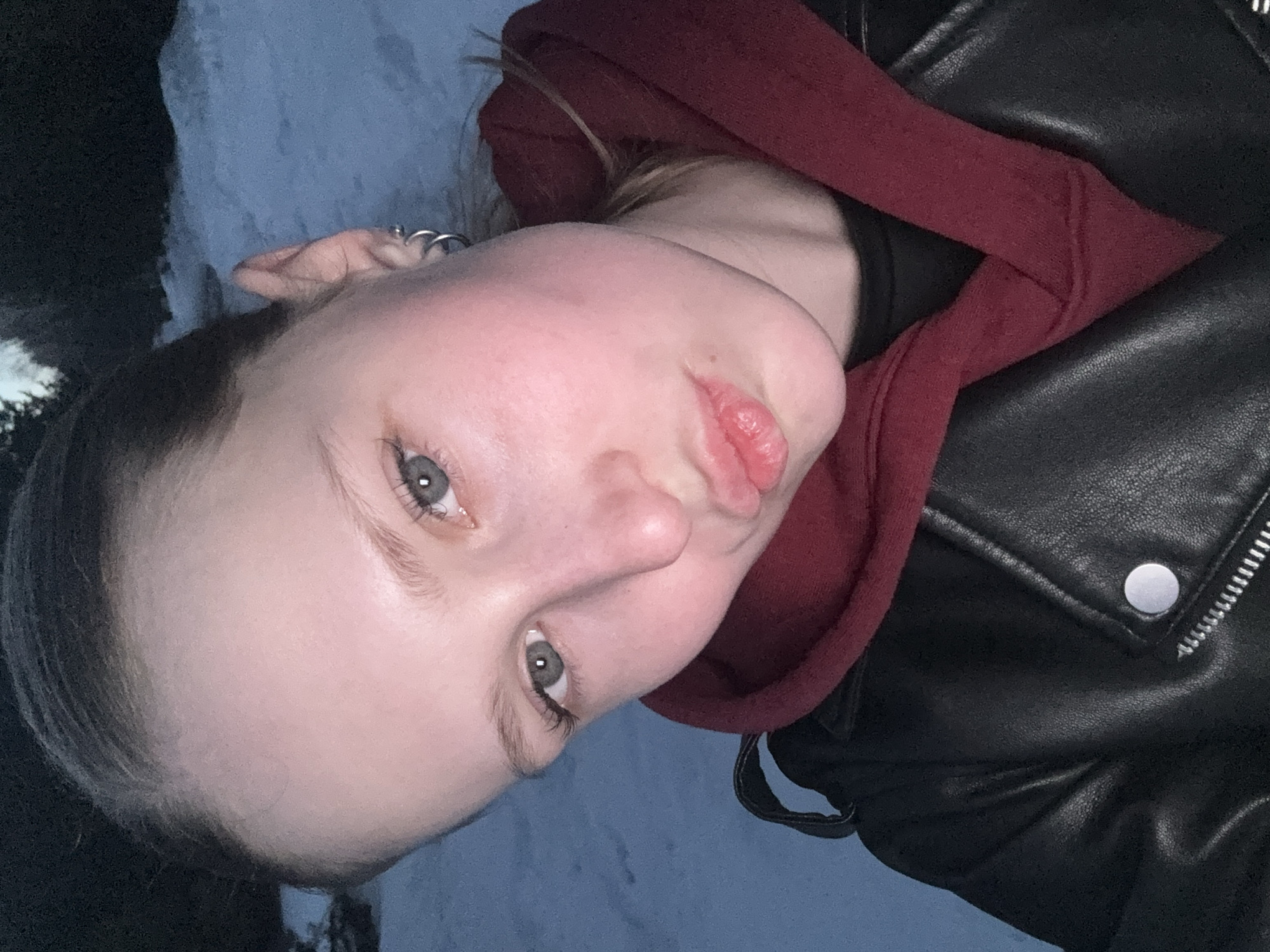 A fair-skinned person with light eyes is positioned in the center, tilting their head to the right. They are wearing a red hoodie and a black leather jacket. An earring is visible on their left ear. The background appears to be a muted blue or grey, possibly depicting an outdoor setting at night. The location is Chaikovsky, Russia.