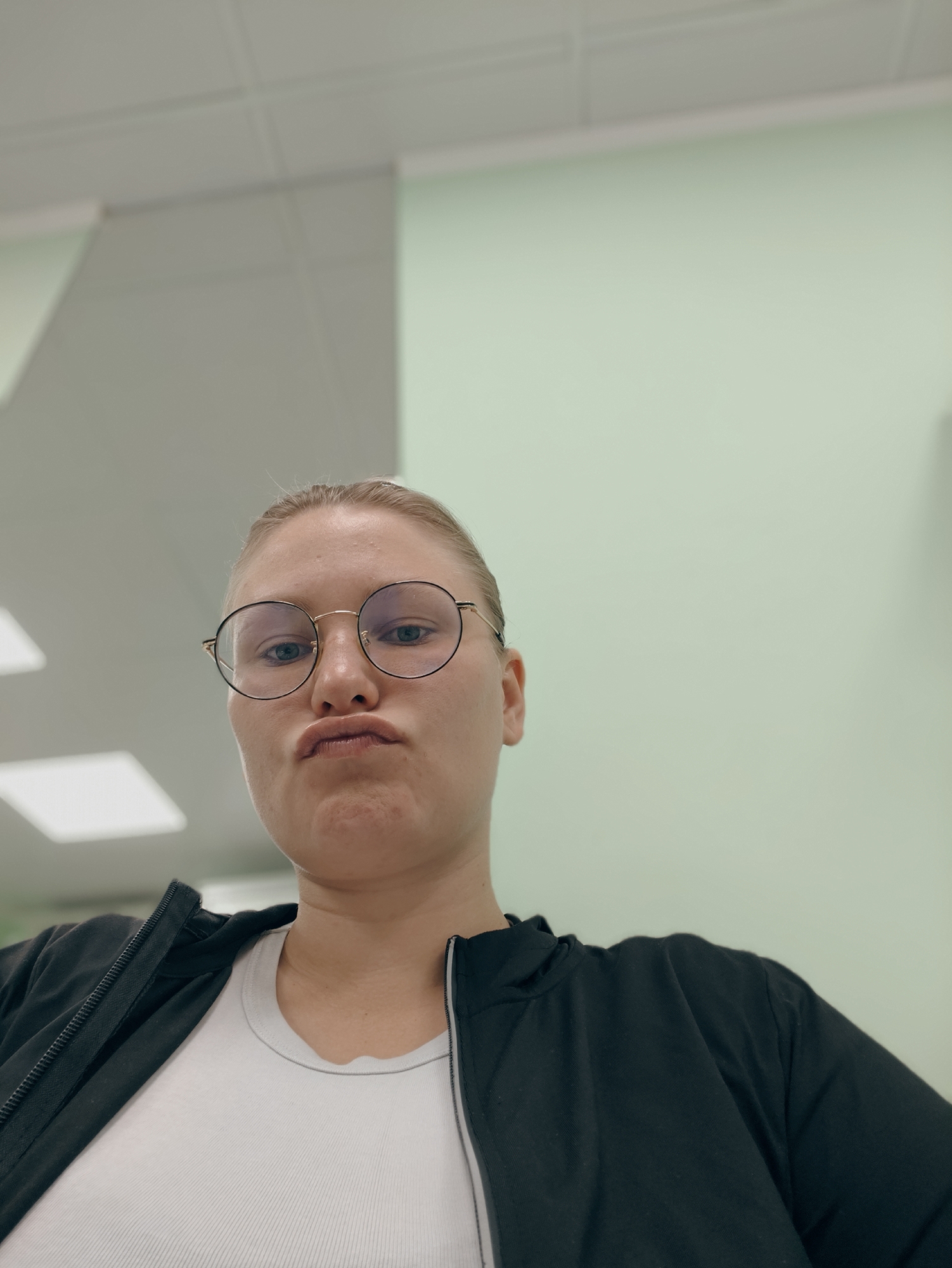 A woman with blonde hair pulled back and wearing round glasses makes a puckered face while looking at the camera. She is wearing a white t-shirt under a black zip-up jacket. The background appears to be an indoor environment with light green and gray walls, and a tiled ceiling with fluorescent lighting. The location is not specified as Podolsk, Russia, nor is there any information to suggest this.