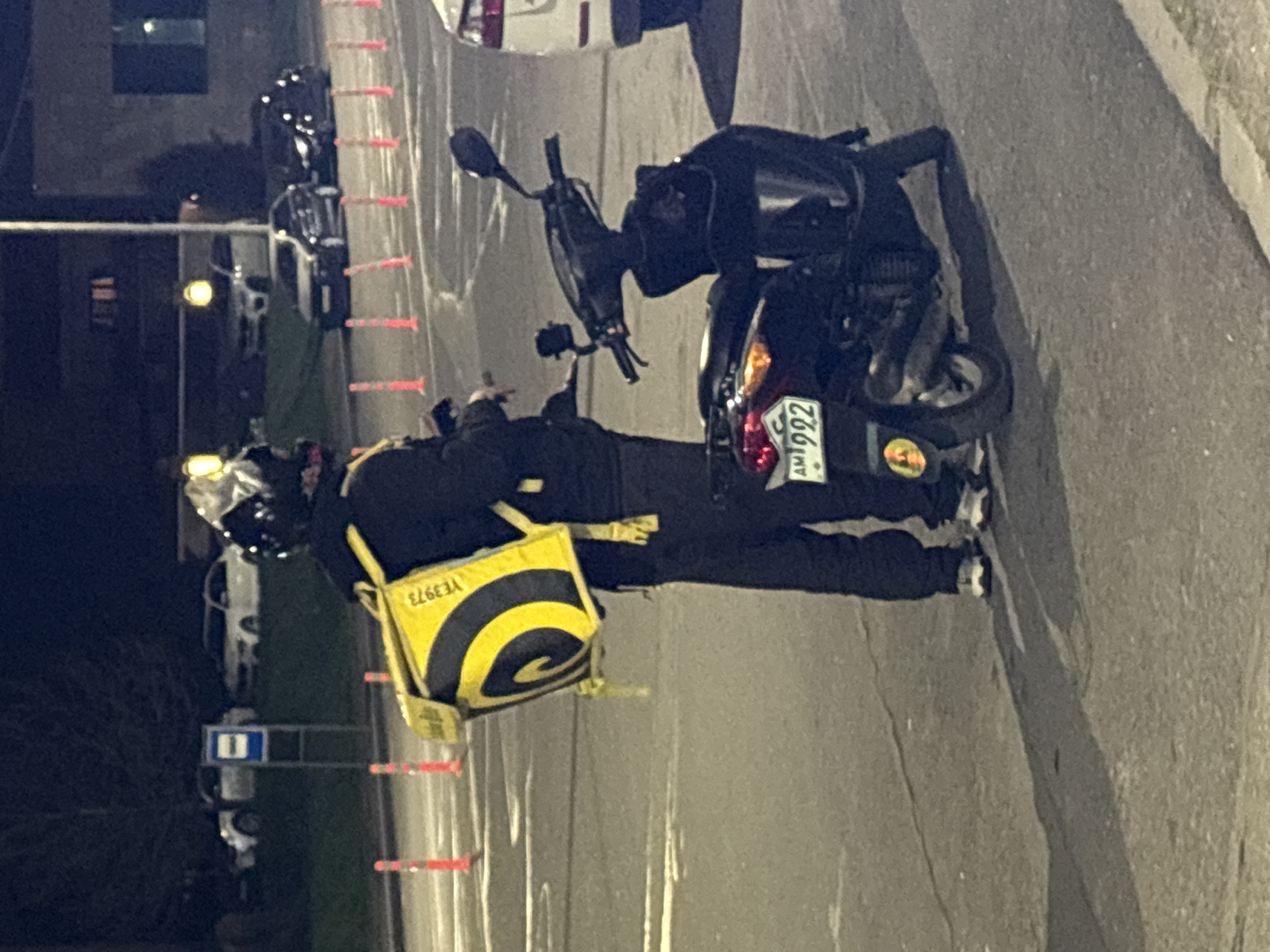 An urban street scene is captured at night, with a delivery rider and their scooter as the central subjects. The image is oriented sideways, with the street running vertically.
A person wearing dark clothing (jacket and pants) and a helmet is standing beside a black scooter. They have a prominent yellow and black delivery bag on their back, featuring a distinctive black spiral logo and the inverted text "YE3973". The scooter is black, with its taillights illuminated. A license plate, clearly visible on the rear of the scooter, displays "AM 992". Below the license plate, a yellow circular sticker or emblem is present.
The setting appears to be a paved street or parking area. In the background, other parked vehicles and dimly lit buildings can be seen. Artificial light sources, likely streetlights, illuminate the area, casting shadows. Red and white striped barriers or poles line what appears to be the edge of the road or a construction zone. Further in the background, a blue rectangular sign with white markings is visible.
The time of day is night, indicated by the darkness and the use of artificial lighting. The activity suggests a momentary pause by a delivery driver, either preparing for a delivery or having just completed one. The "AM" on the license plate confirms the location as Armenia, aligning with the provided context of Yerevan.