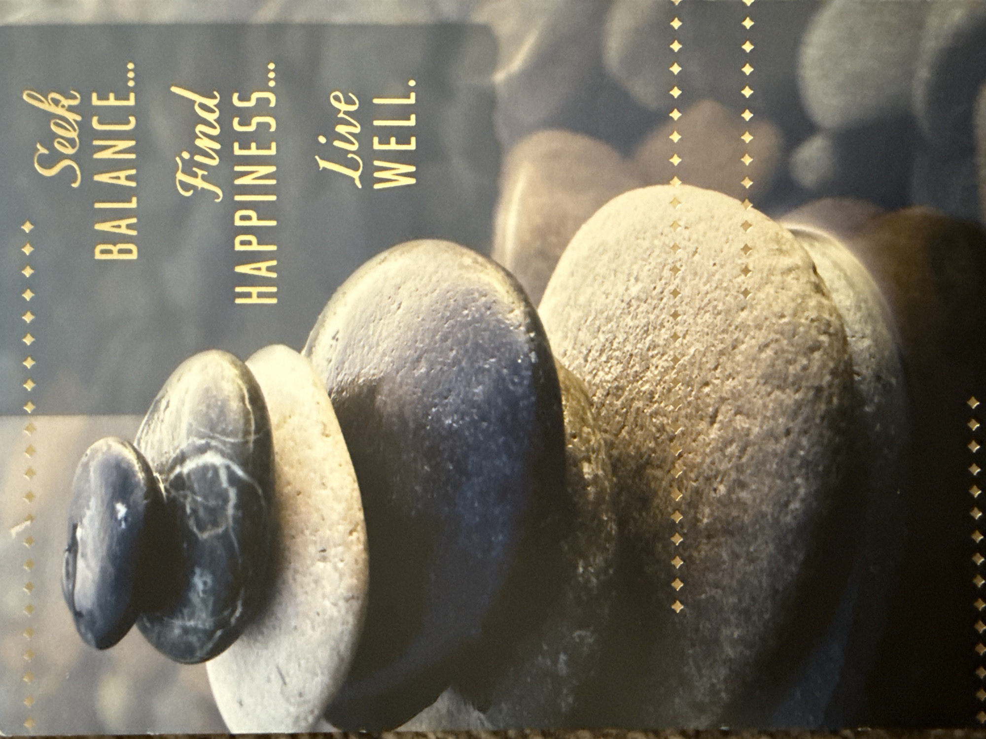 The image is a close-up photograph of stacked stones with text overlay. The stones are smooth and rounded, with varying colors from dark gray to light tan. Some of the stones have visible markings or veins. The text, in a golden, cursive and block font, reads "Seek BALANCE... Find HAPPINESS... Live WELL." The background appears to be a blurred underwater scene with more stones. The overall impression is one of tranquility and mindfulness. The image is a postcard or greeting card and does not depict any specific location in West Sacramento, United States of America.
