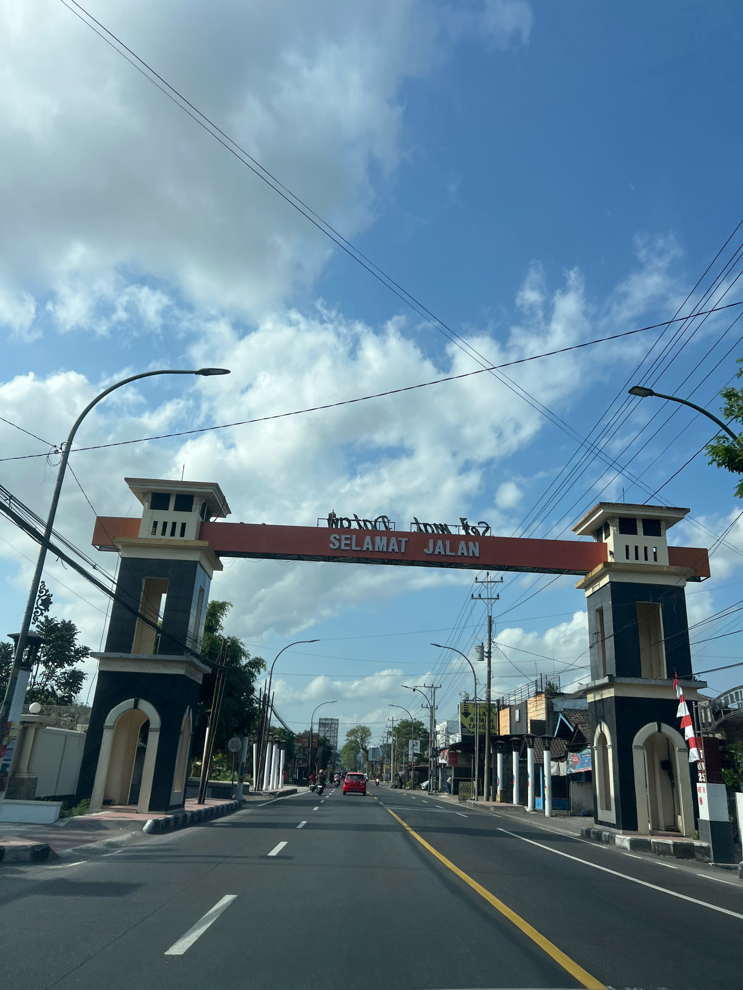 The image shows a wide, paved road in the city of Payaman, Indonesia, under a bright blue sky with scattered clouds. An archway spans the road, featuring two dark pillars with orange accents and a central orange banner displaying the Indonesian phrase "SELAMAT JALAN" in white lettering. The phrase translates to "Have a good trip."
On either side of the road, there are sidewalks and buildings, some of which appear to be shops or residences. Streetlights are visible along the road. A red car is driving away from the viewer on the right side of the road, and a few motorcycles are also present. The overall environment suggests a typical day in a populated area.