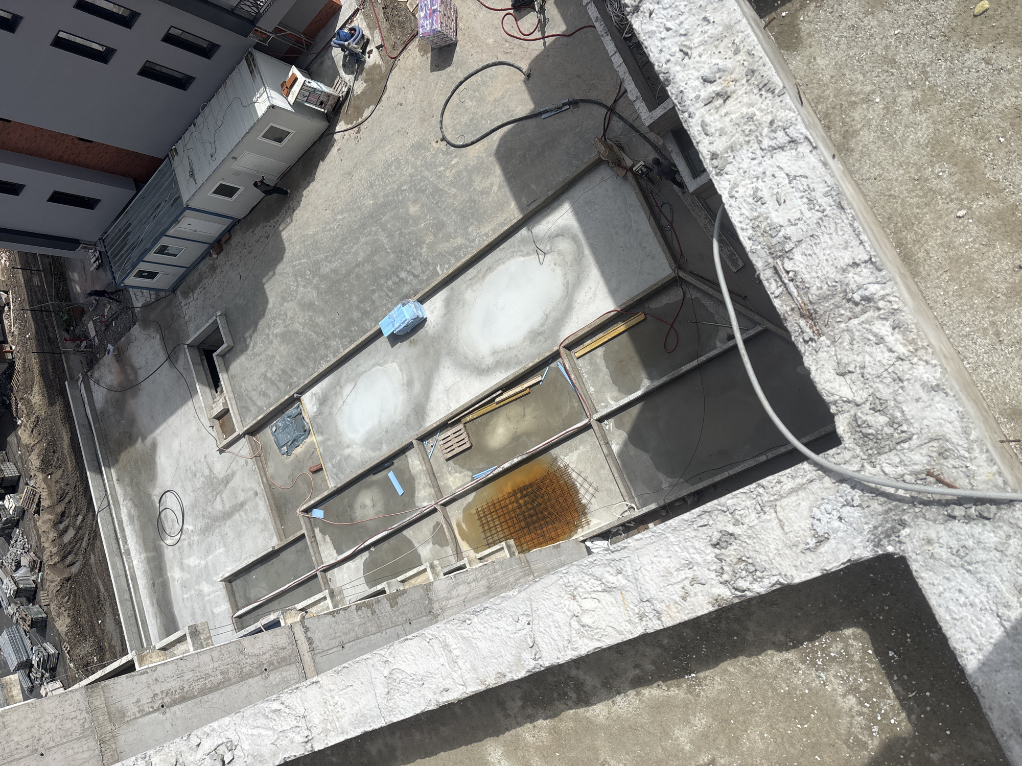 The image shows an elevated view of a construction site in Burgas, Bulgaria. A multi-story building is partially visible to the left. A modular structure, likely a construction office, is positioned adjacent to the building. The ground level reveals a concrete structure divided into rectangular sections, some containing water or construction debris. Reinforcement mesh is visible in one section. Construction materials and equipment are scattered around the site. Concrete support beams are in the foreground, suggesting the viewpoint is from an unfinished upper level.
