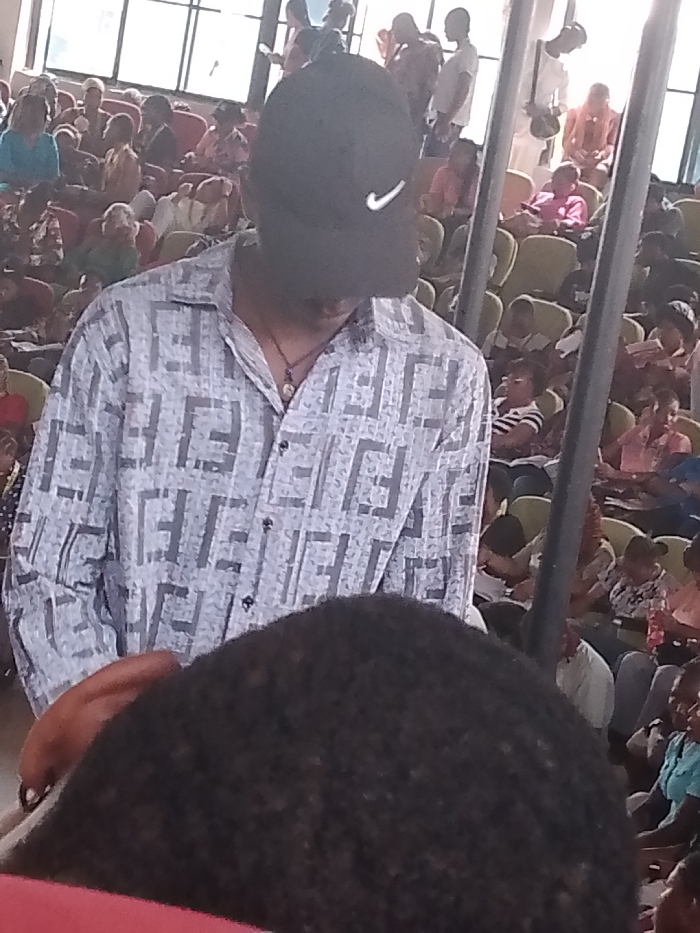 A man wearing a black baseball cap with a white Nike logo and a light blue button-down shirt with a gray Fendi logo pattern stands out in a densely populated interior. The man has a thin necklace and is situated in front of an audience seated in rows of chairs. The setting appears to be a large indoor space, possibly a stadium or auditorium, with a mix of people of all ages. The photograph was taken in Umueshi, Nigeria.