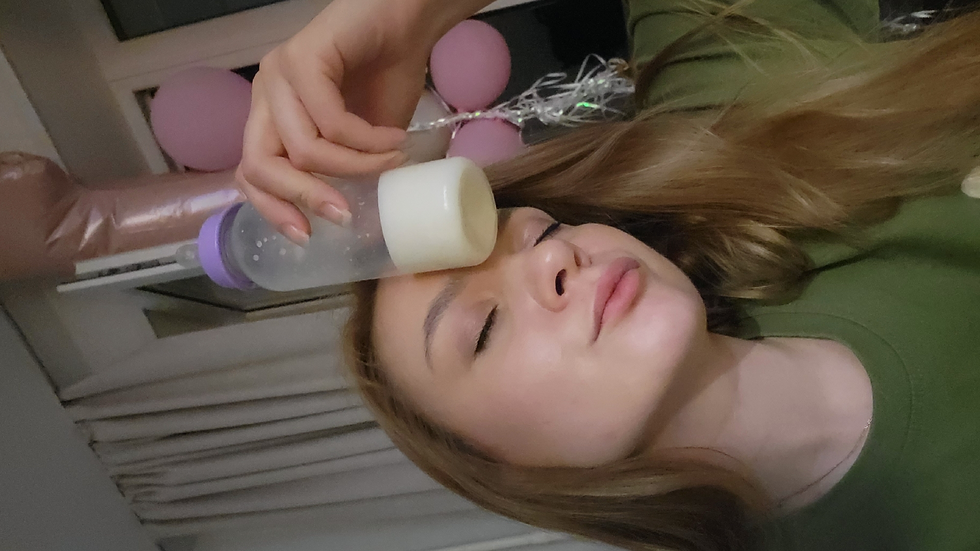 A person, presumably female, is lying on their side with a baby bottle being held against their forehead. The bottle is translucent with a purple nipple and a white cap. Light pink balloons and some sort of shiny garland are in the background. The person has fair skin and long, light brown hair. They are wearing a green shirt. Based on prior knowledge, the image was taken in the city of Chaikovsky, Russia.