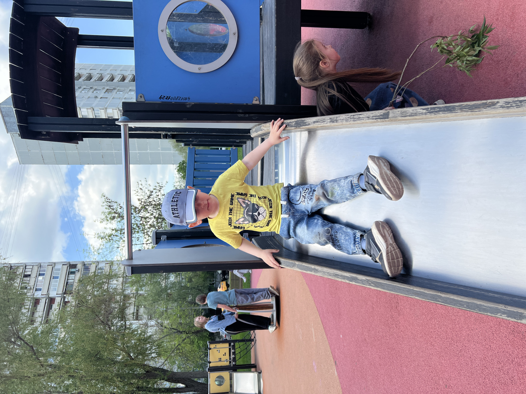 A boy is on a slide on a playground with a red surface. He is wearing a yellow t-shirt, blue jeans, a gray baseball cap, and sneakers. A girl is looking up, sitting near a blue structure with a circular window, holding a branch with green leaves. Behind the slide, two people are standing near playground equipment, with a residential building visible in the background. The location is Moscow, Russia.
