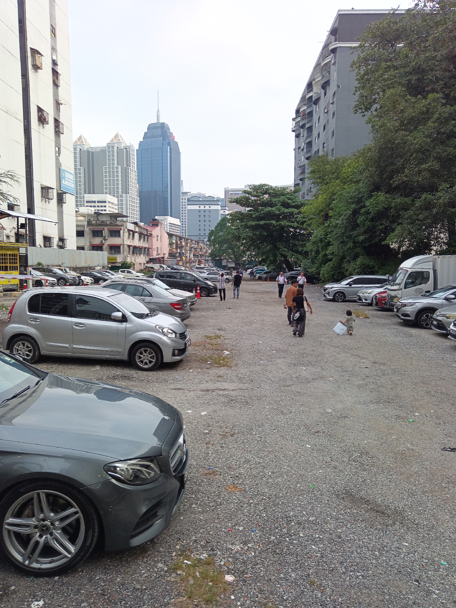 The image depicts a dusty, gravel parking lot in Kuala Lumpur, Malaysia, with several cars parked. In the foreground, the front right portion of a dark gray Mercedes-Benz is visible. Further into the lot, a silver Perodua Myvi is parked, and behind it are other vehicles including a silver Honda HR-V and a white Isuzu truck. A group of people, including two men in orange shirts, one person in a brown shirt, and a woman in black, are walking in the parking lot. A small child in a light-colored dress is also seen walking away from the camera, holding a white object. In the background, various buildings are visible, including a tall blue skyscraper, residential buildings, and trees. The sky is overcast.