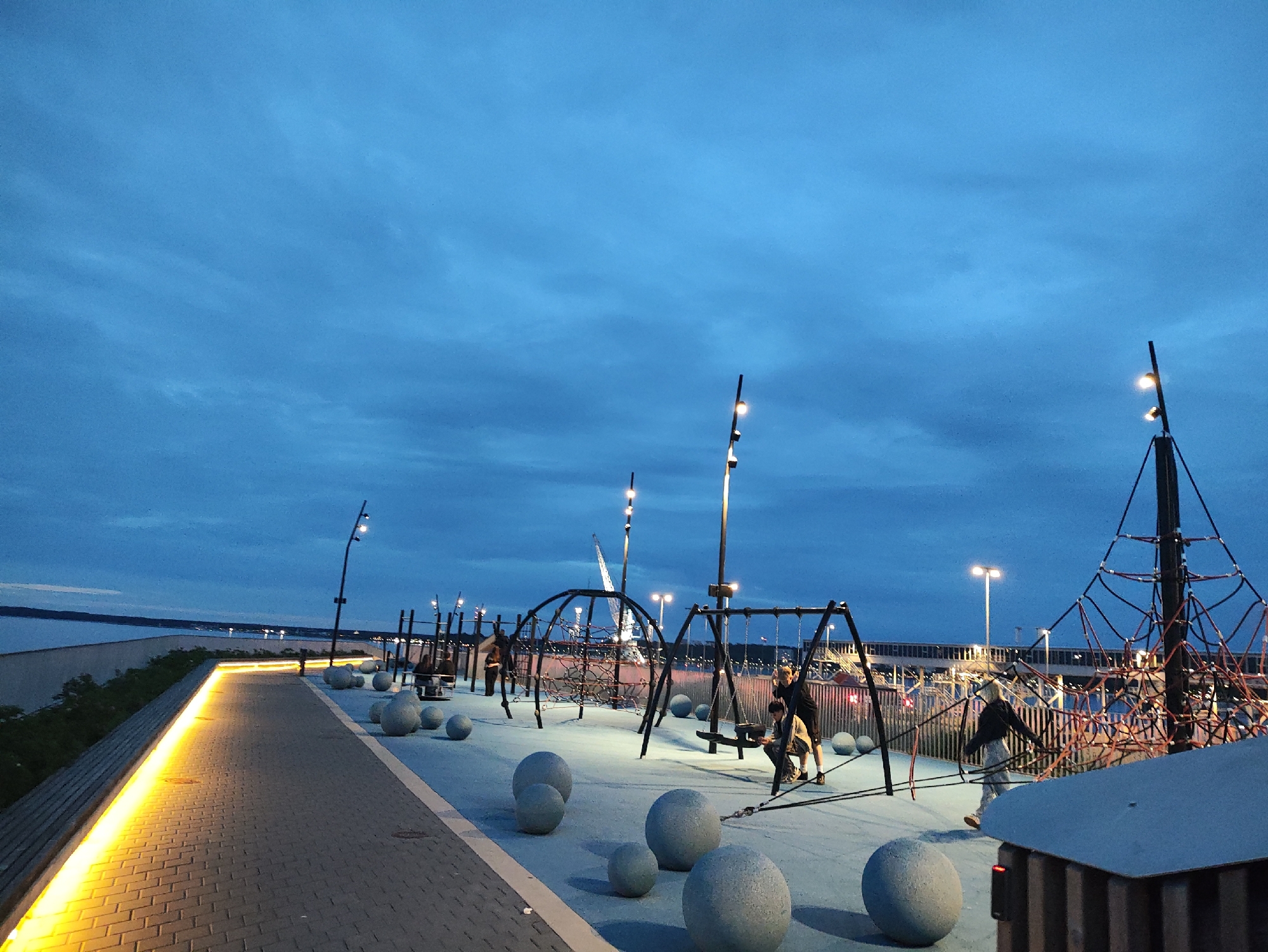 The image depicts a public recreational area at dusk, likely in the city of Tallinn, Estonia, possibly near the waterfront. The scene features a paved walkway illuminated by a continuous band of warm yellow light along its edge. Beyond the walkway, a blue-toned playground area is visible. This area contains various play structures, including swings, a climbing net, and horizontal bars. Several large, spherical stone objects are scattered across the playground surface. A few individuals are present, with two people actively engaged with a swing set, and others appearing to be walking or observing. In the background, the calm water of a bay or sea stretches towards the horizon, with distant lights of a cityscape or port visible. The sky above is a deep blue with scattered clouds, indicating twilight conditions.