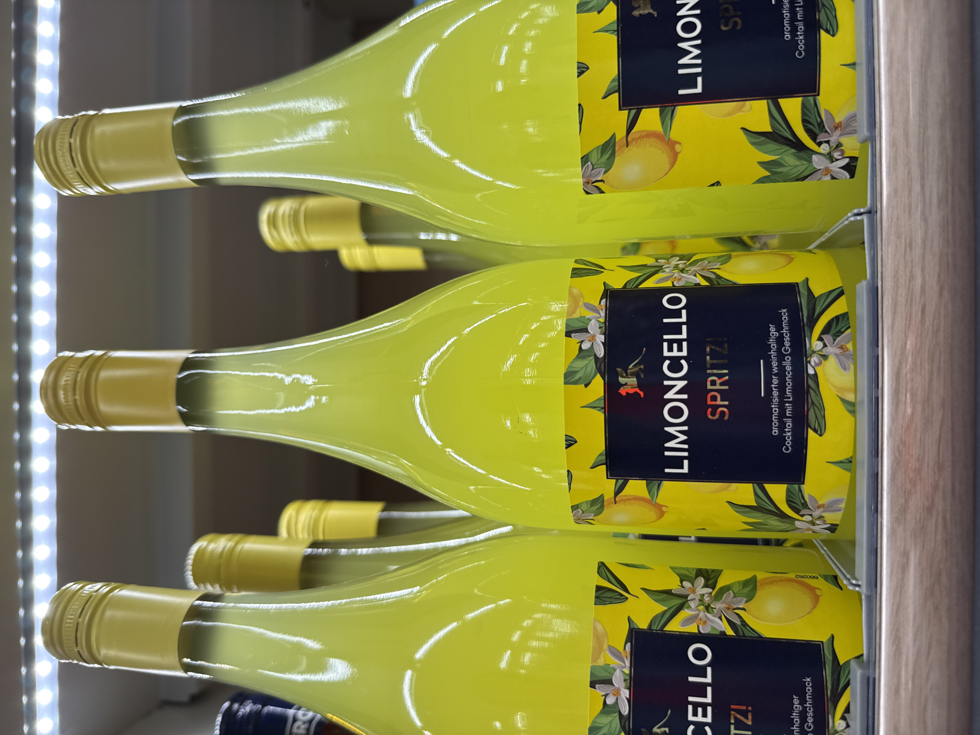 This image shows a close-up, eye-level, slightly low-angle view of several bottles of Limoncello Spritz arranged on a shelf in a retail environment, likely a liquor store or supermarket. The bottles are tall and slender with bright yellow caps and a vibrant yellow liquid inside. Each bottle has a prominent label featuring a yellow background with illustrations of lemons, lemon blossoms, and green leaves. Text on the labels includes "LIMONCELLO SPRITZ!" and "aromatisierter weinhaltiger Cocktail mit Limoncello Geschmack."
The lighting appears to be artificial, possibly from LED strips illuminating the shelf from above, creating reflections on the glass bottles. In the background, other bottles and shelves are visible but out of focus. The overall impression is a product display, clean and organized. The location in the city of Bad Lauterberg im Harz, Germany, is not explicitly depicted in the image itself, but this description is provided as context.