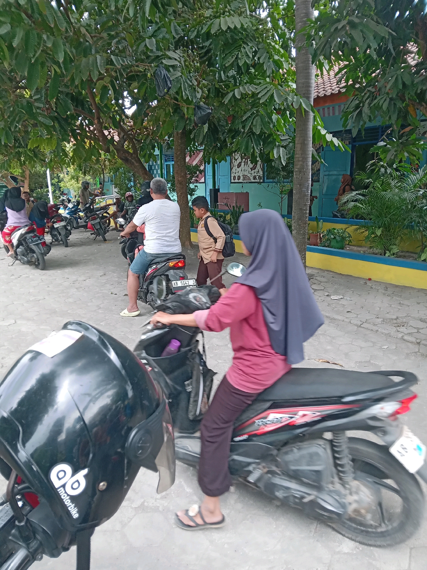 The image depicts a scene outside a building with a turquoise and orange facade, likely a school or community center, in Sitimulyo, Indonesia. Several people are present, mostly on or around motorcycles. In the foreground, a black motorcycle helmet with a "qb motorbike" logo is prominently displayed. A woman wearing a dark purple hijab and a pink shirt is seated on a black motorcycle in the mid-ground, facing right and her hand on the handlebar. To her left, a man in a white t-shirt and blue shorts is on another motorcycle, facing left, with his right leg extended. Behind him, a young boy in a light brown and tan school uniform with a backpack walks away from the motorcycles. Several other motorcycles and people are visible in the background, suggesting a drop-off or pick-up situation. Trees with abundant green leaves provide shade over the paved area where the motorcycles are parked. The ground appears to be a mix of concrete and possibly some gravel. The overall atmosphere seems to be one of everyday activity.