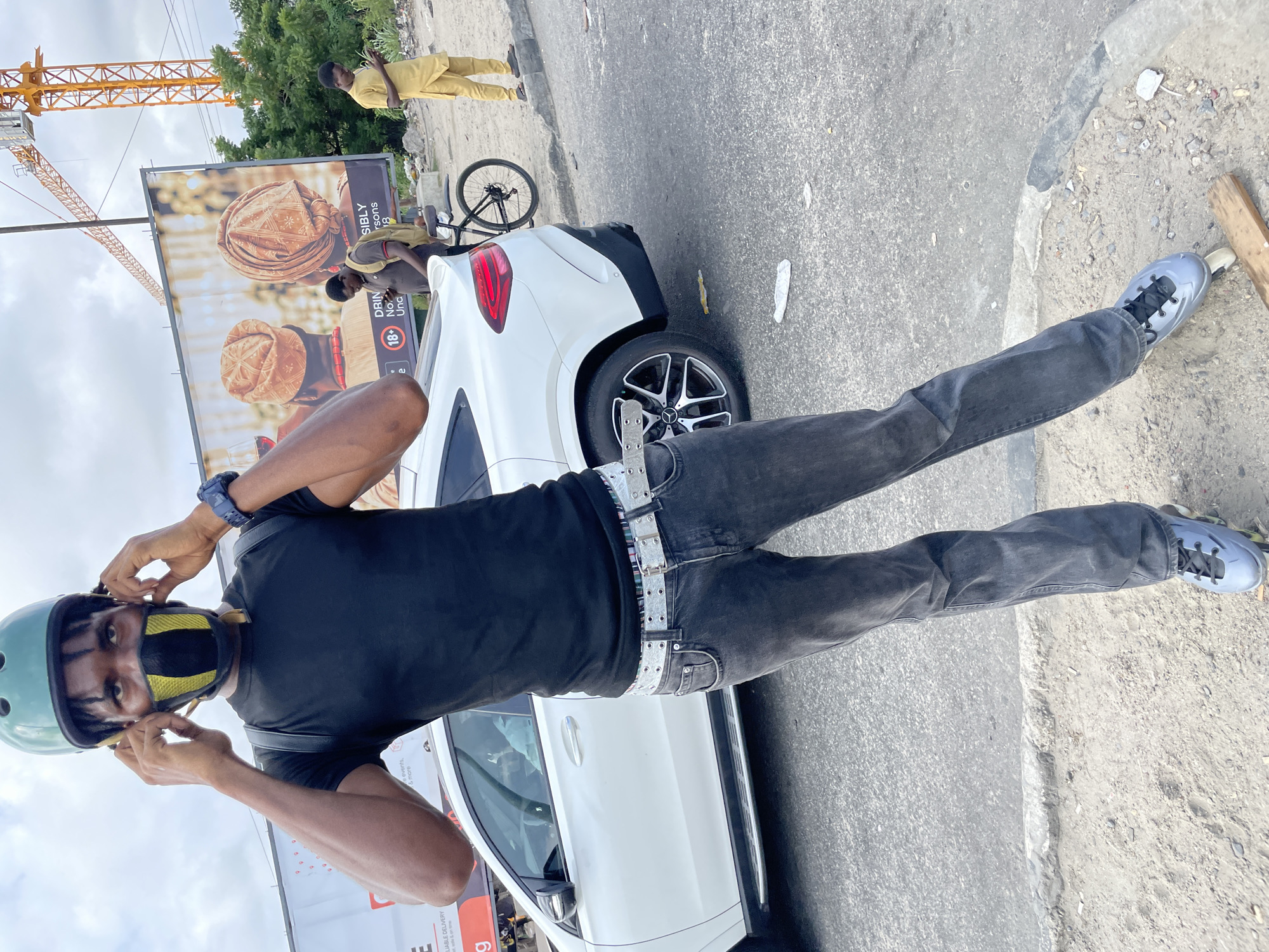 In Ilado, Nigeria, a young Black man wearing a black t-shirt, grey jeans, roller skates, and a light blue helmet, is adjusting a face mask with black and yellow detailing. He has a black backpack on. Behind him, a white SUV is parked next to a billboard featuring two people in traditional headwear. Two other Black men are visible: one wearing a yellow outfit on the right, and another in a brown shirt and backpack standing near a bicycle. A yellow construction crane is also visible in the background against a cloudy sky. The ground is a mix of asphalt and sand, with some debris scattered around.
