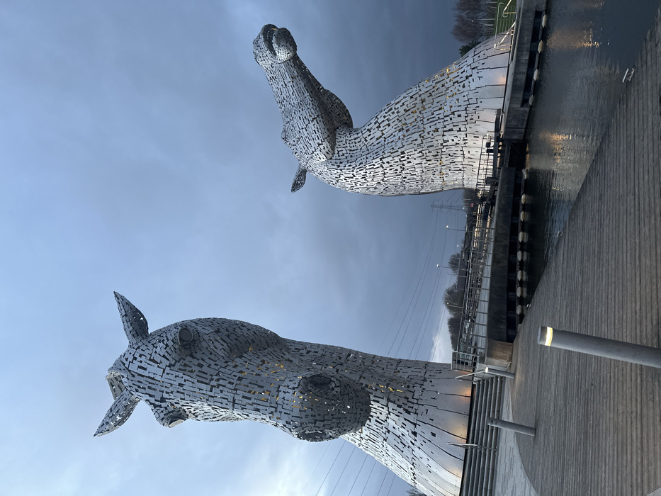 The image displays a wide-angle view of The Kelpies, two colossal equine head sculptures, situated at The Helix park in Falkirk, United Kingdom, under an overcast sky.
In the foreground, a wooden boardwalk with visible plank lines extends from the lower left towards the center-right. Three cylindrical grey bollard lights with illuminated white tops are visible on the boardwalk. To the right of the boardwalk, a calm body of water, likely a canal, reflects ambient light and portions of the structures.
The dominant subjects are the two metallic horse head sculptures. The sculpture on the left is larger and positioned closer to the viewer, facing slightly left and upwards. Its surface is composed of numerous interlocking, reflective metal plates, with internal illumination visible through the gaps, highlighting its eye sockets and nostril area. A set of concrete stairs with metal handrails ascends to the base of this sculpture from the left.
The sculpture on the right is positioned further back and appears smaller due to perspective. It depicts a horse's head raised and looking towards the right. Its construction mirrors the left sculpture, featuring segmented metal plates and visible internal lighting.
Between the boardwalk and the sculptures, a concrete or paved area is visible, with a bridge structure extending across the water, partially obscured by the sculptures. In the distant background, leafless trees line the horizon, and utility poles or electrical pylons are discernible against the sky. The sky above is largely grey and cloudy, darker towards the upper right, indicating low light conditions, possibly dusk or dawn. No individuals are present within the frame.