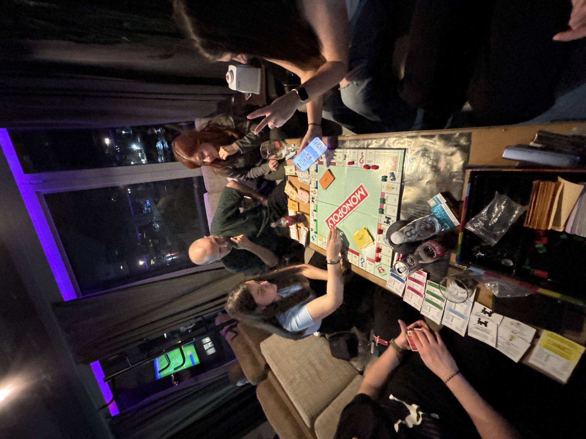 A group of people are playing Monopoly around a wooden table indoors. The game board is visible, along with Monopoly money, property cards, and game pieces. A bald man is at the left, a woman with red hair is sitting to his right. Two women are standing in the upper right corner, one is using a smartphone to take a picture. The lighting is subdued, with a window showing a cityscape at night in the background. A sofa is behind the table with a women seated on it with the properties in front of her. The city of Plovdiv, Bulgaria cannot be confirmed from the image.