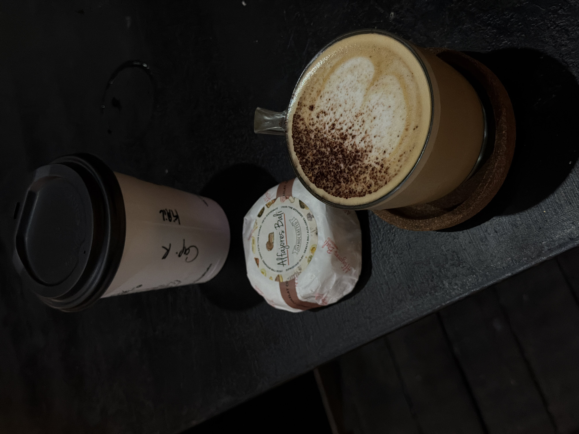 A close-up, overhead shot shows a coffee cup and a wrapped pastry on a dark, textured surface. The coffee cup, made of clear glass with a handle, contains a latte with foamed milk art resembling a heart, dusted with cocoa powder. It rests on a woven coaster. To the left of the coffee, a white paper cup with a black lid is partially visible, with handwritten text on its side that appears to say "Cap. R" and "Khil". Between the two cups, a round pastry, wrapped in white paper, is adorned with colorful illustrations and text that reads "Alfajores Bali HANDCRAFTED". The overall scene suggests a casual moment, likely in the morning or afternoon, given the warm tones of the coffee and pastry. The dark background and the way light casts shadows hint at an indoor setting with subdued lighting. The location context, North Kuta, Indonesia, suggests a tropical setting, though no specific environmental cues are visible. The focus is on the food and drink items, creating an inviting still life.