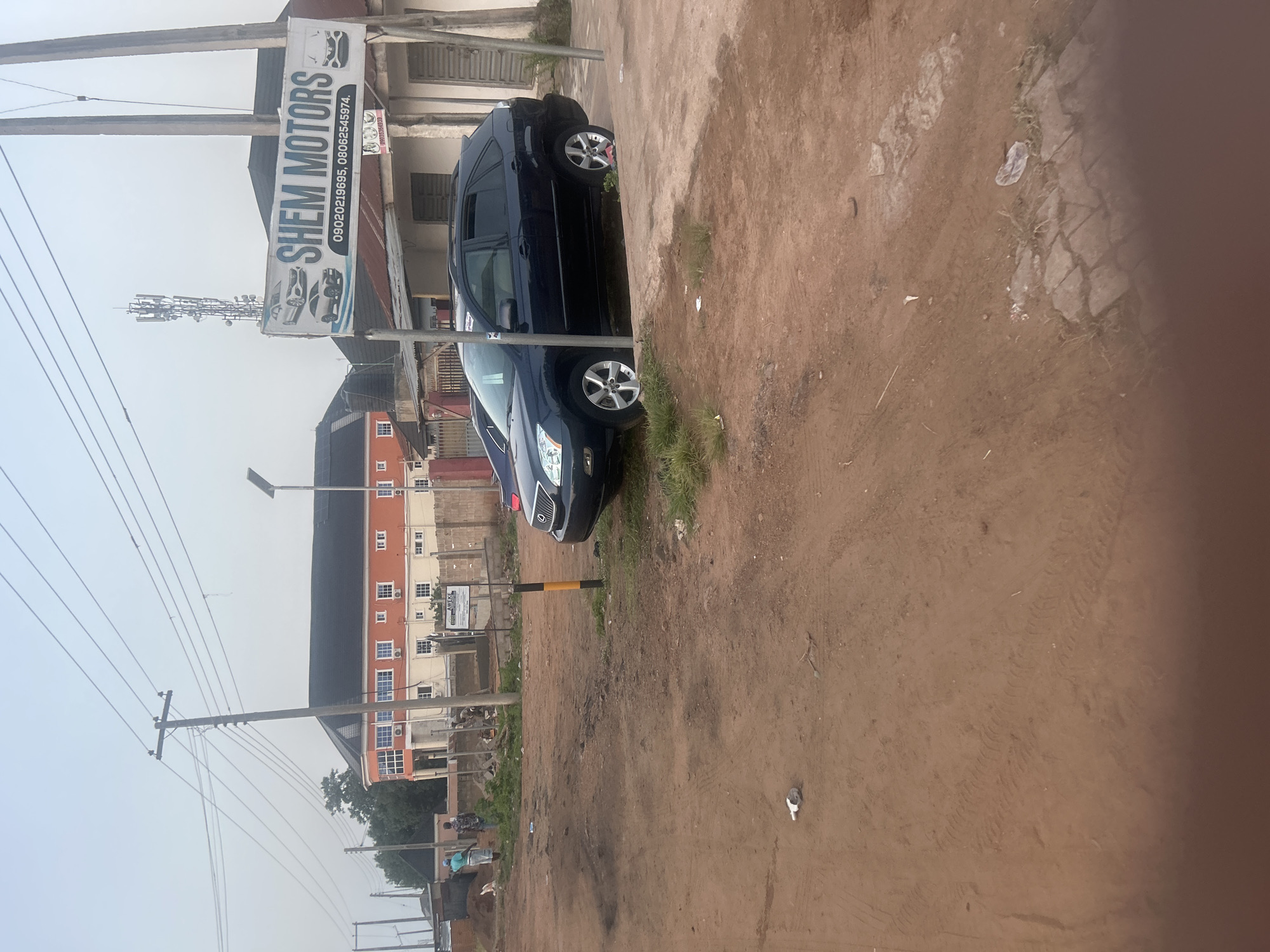 The image depicts a street scene in Ogbomosho, Nigeria, likely in a commercial area. In the foreground, a dark blue SUV is parked on a dirt lot. The vehicle appears to be a Lexus RX. Behind the SUV, a building with an orange facade and white-trimmed windows is visible, suggesting it could be a business or residential complex. A prominent sign for "SHEM MOTORS" with contact numbers is affixed to a pole in front of a building to the right of the orange structure. This suggests a car dealership or repair shop. Several telephone poles and power lines are present, indicating urban infrastructure. Two individuals are walking in the background near the orange building, and construction activity appears to be taking place in front of it. The overall environment is dusty with an unpaved road surface.