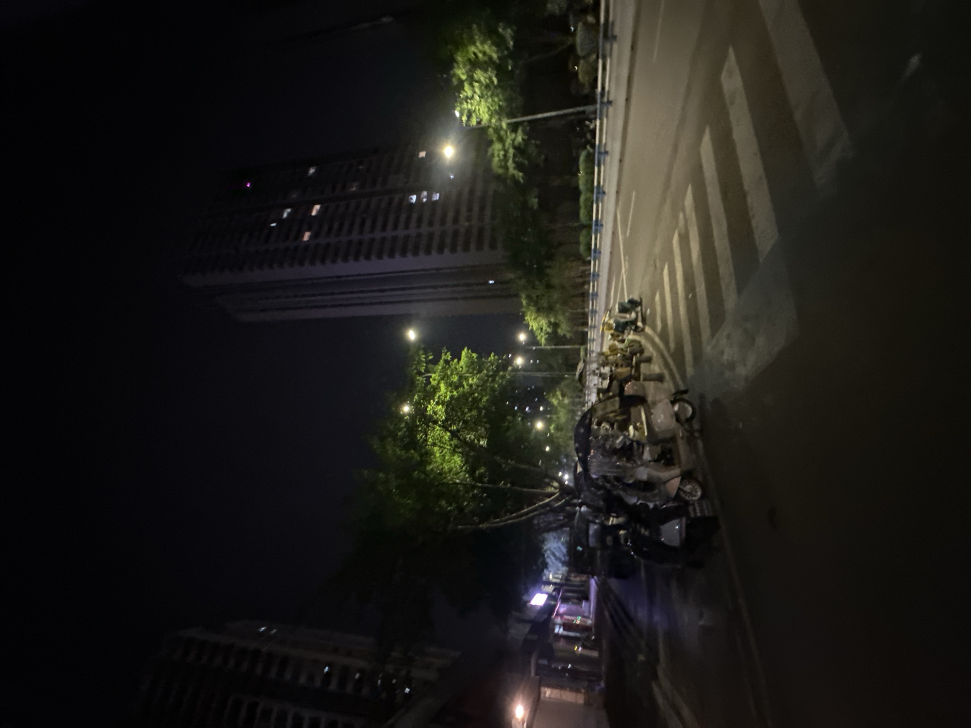 The image shows a nighttime scene on a street in Chongqing High-tech Zone, China. A tall, modern building is visible in the background, illuminated by interior lights. In the foreground, several parked motorcycles and scooters are lined up along the sidewalk. The street is paved, with a pedestrian crossing marked by white stripes. Trees with green foliage are present along the side of the road, and streetlights cast pools of light. The overall atmosphere is urban and active, despite the late hour.