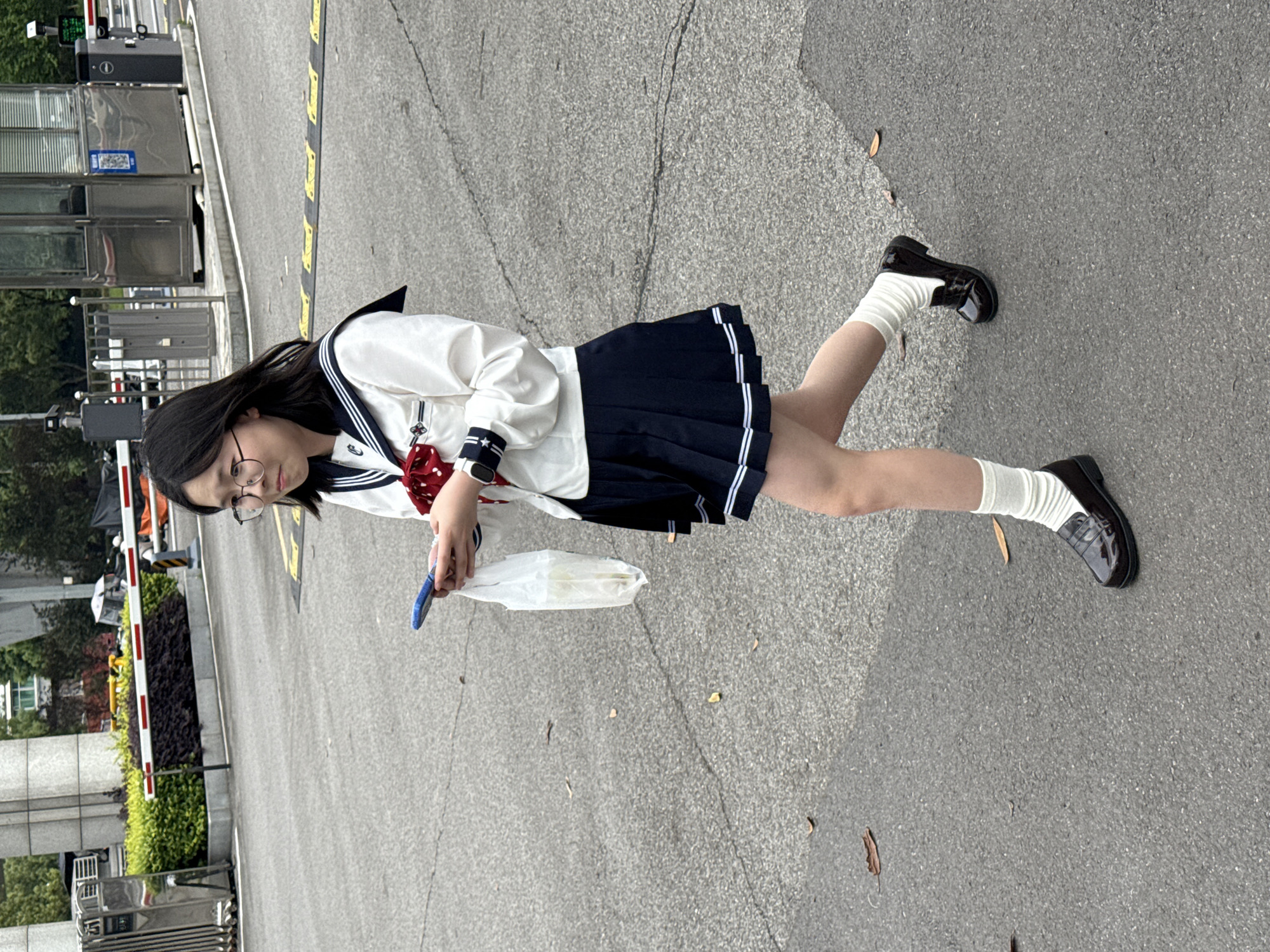 A young woman stands on a paved surface, likely a road or parking lot, in an outdoor setting. She wears a white sailor-style blouse with a navy skirt that has white stripes near the hem, white socks, and black loafer-type shoes. Her outfit resembles a Japanese school uniform. She is also wearing round glasses and has dark hair. She is holding a mobile phone in one hand and a white plastic bag in the other.
The background includes foliage, a security gate, and what appear to be metal security barriers. Based on these features, the location may be near a controlled entrance to a building or area. It appears to be in the city of Jiangjin in China.