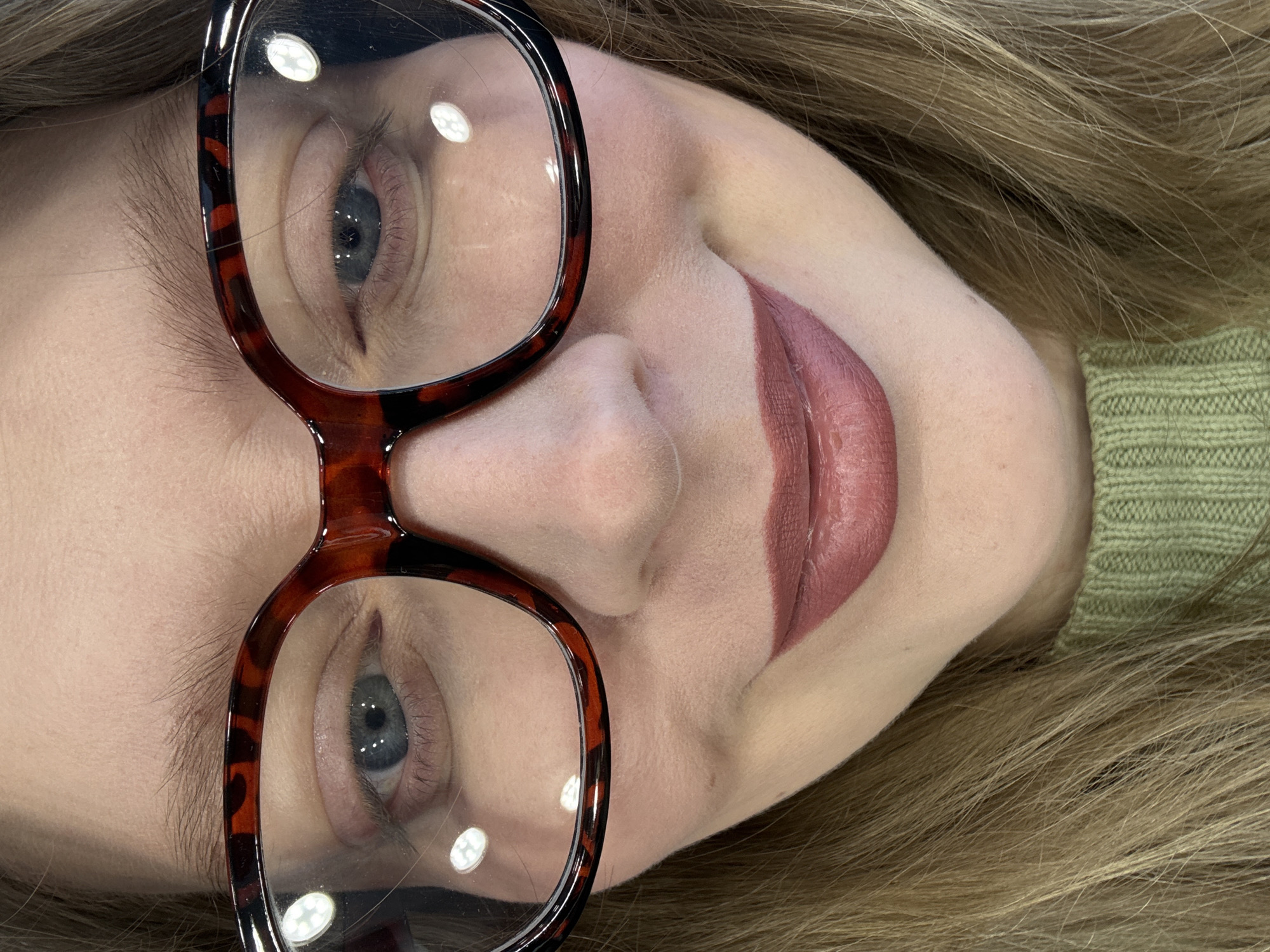 A close-up, tilted portrait shows a fair-skinned woman wearing glasses. Her eyes are blue-grey, and her lips are painted a muted mauve color. The glasses have tortoise-shell patterned frames. She has light brown hair. A green sweater or shirt is visible on the right side of the image. The location is not discernible from the image.