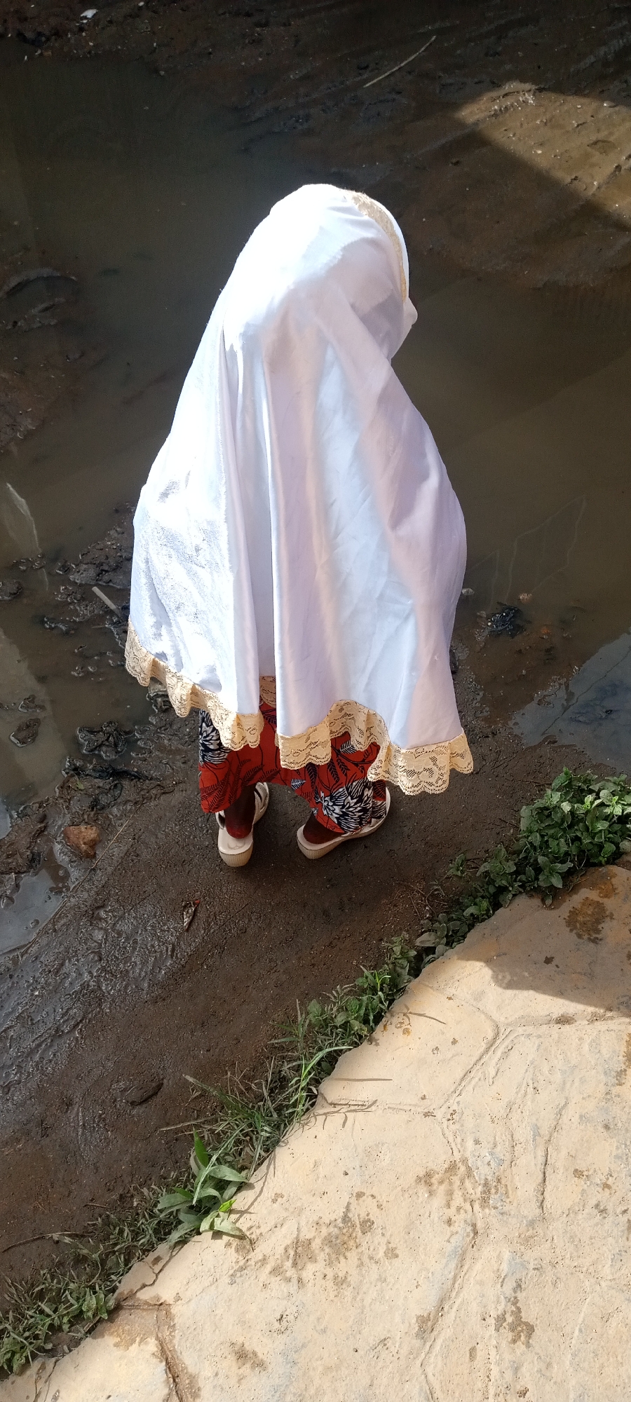 A person wearing a white garment with lace trim and a patterned orange and red garment stands on a muddy bank in Naraguta Hausawa, Nigeria. The person faces away from the viewer, with a body of water to the left and vegetation along the bank. The person is wearing brown sandals. To the right, is a concrete surface.