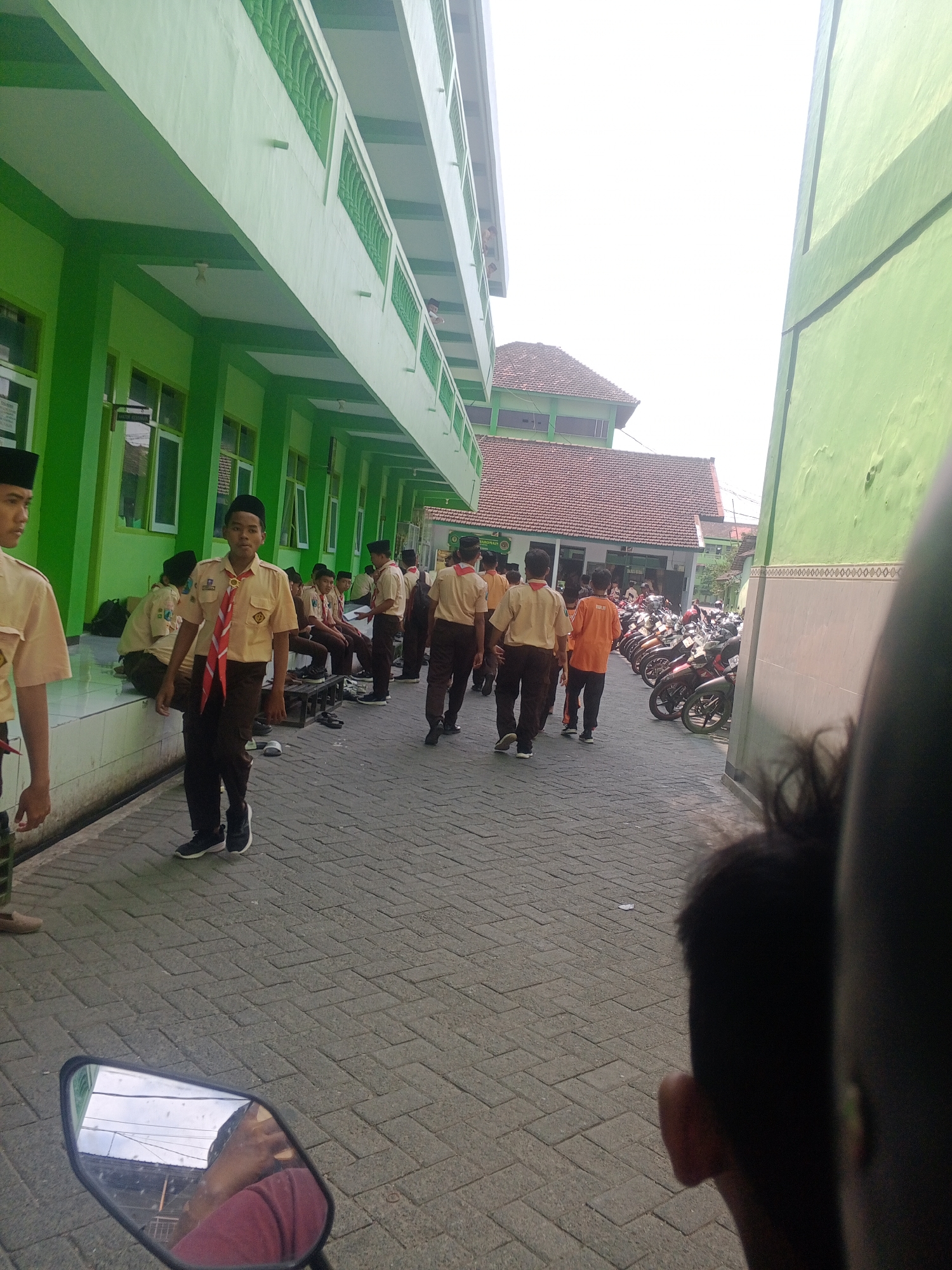 The image depicts an outdoor scene at a school, likely in Ronowijayan, Indonesia, given the architecture and the presence of students in uniform. The dominant colors are green from the school building's facade and tiled walkways. Several students, identifiable by their light brown shirts, dark pants, and dark peci hats, are present. Some are walking, while others are seated or standing in small groups along the walkway. A row of motorcycles is parked to the right of the walkway, adjacent to a light gray wall. The sky above is bright and appears to be a clear, sunny day. In the foreground, a motorcycle rearview mirror reflects a portion of the scene, including what appears to be the back of a person's head and shoulder.