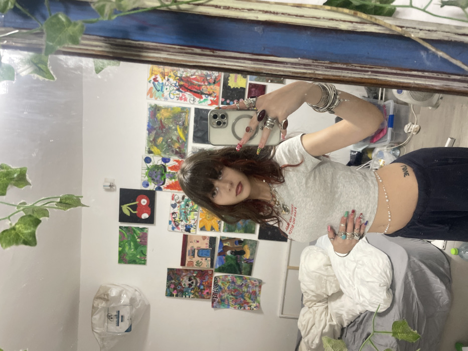 A female individual is depicted taking a mirror selfie within a residential room in Raanana, Israel. The subject has dark, layered hair with reddish undertones and bangs, a light complexion, and dark lipstick. She wears a grey cropped t-shirt with red text (partially visible: "TRUST NO BITCH"), dark blue sweatpants, and a delicate pearl and bead waist chain. Her left hip displays a tattoo spelling "LOVE". Numerous rings adorn her fingers, and multiple silver bracelets are on her left wrist. Her fingernails are long and elaborately decorated. She holds a light-colored smartphone (iPhone) in her right hand, angled towards the mirror, while her left hand rests on her abdomen. The mirror, framed in a dark blue or black material, occupies the right side of the frame.
The wall reflected behind the subject is extensively covered with approximately twenty varied artworks, ranging from abstract designs and a red cherry on a black background to a skull, a house, and a sunflower. To the left, an unmade bed with white and grey bedding is visible. Artificial green ivy vines hang from the ceiling, reflected in the upper portion of the mirror. On the floor, reflected in the lower right, are a white portable fan, a clear plastic storage container with a blue lid, and a green bottle.